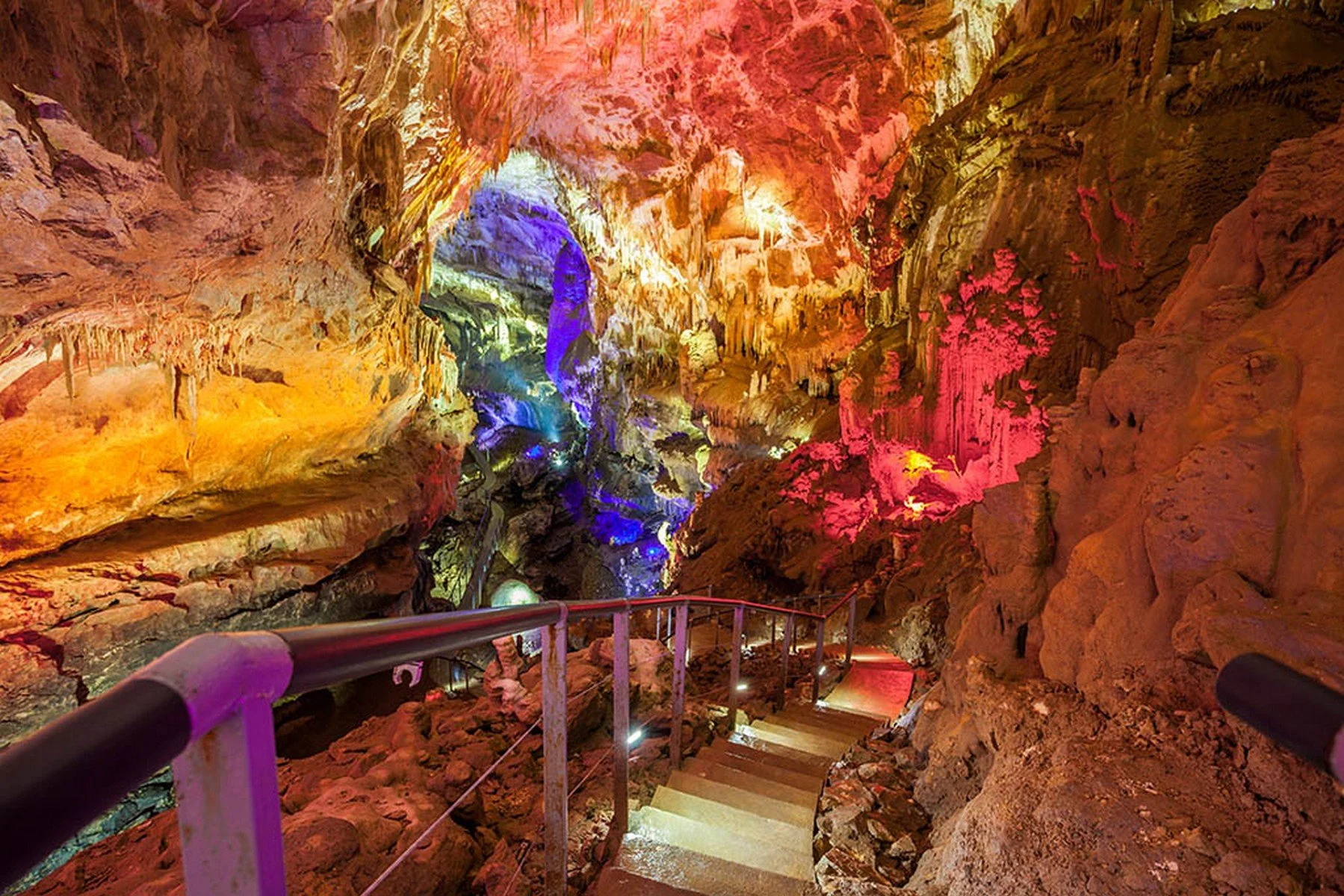 Colorfully illuminated cave with a viewing platform, railing, and a path with stairs leading down into the cave.