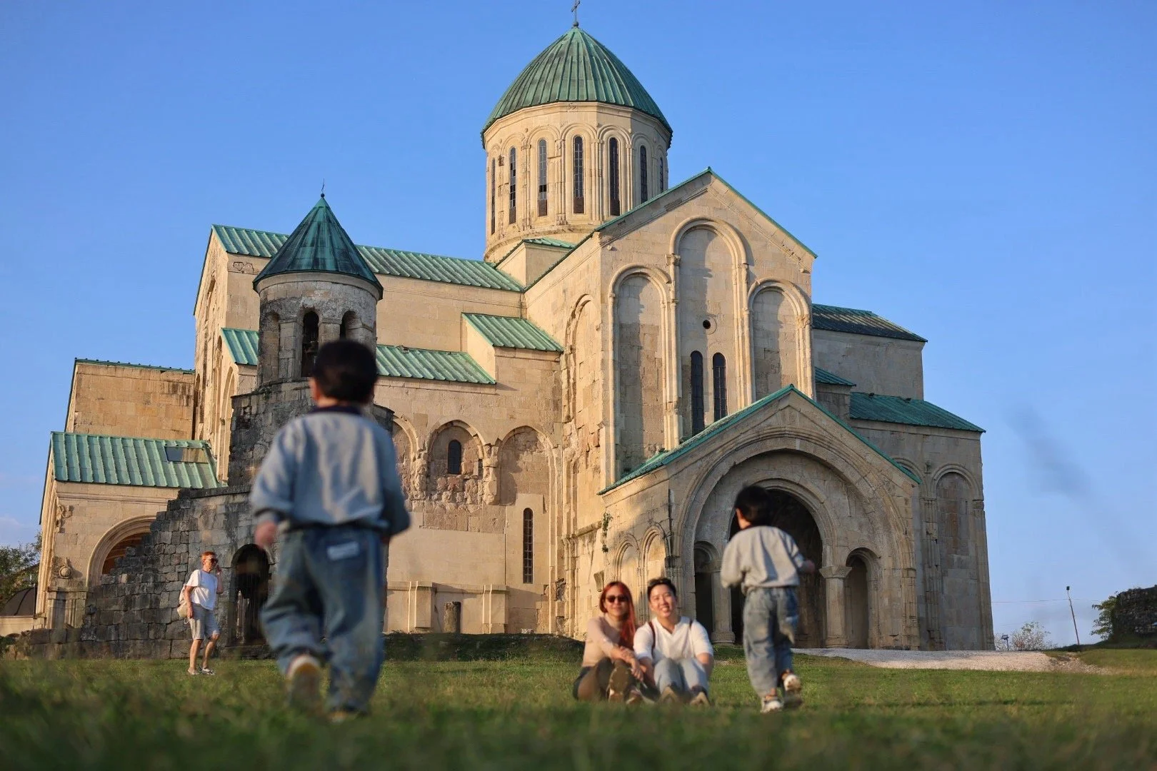 Family sitting on the grass in front of a historic stone church with green roofs and a central dome, under a clear blue sky.