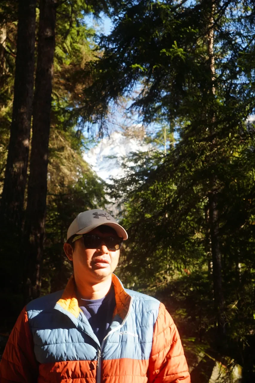 A man wearing sunglasses, a baseball cap, a blue and orange puffy jacket, and a dark shirt, standing outdoors in a forest with tall trees and sunlight filtering through.