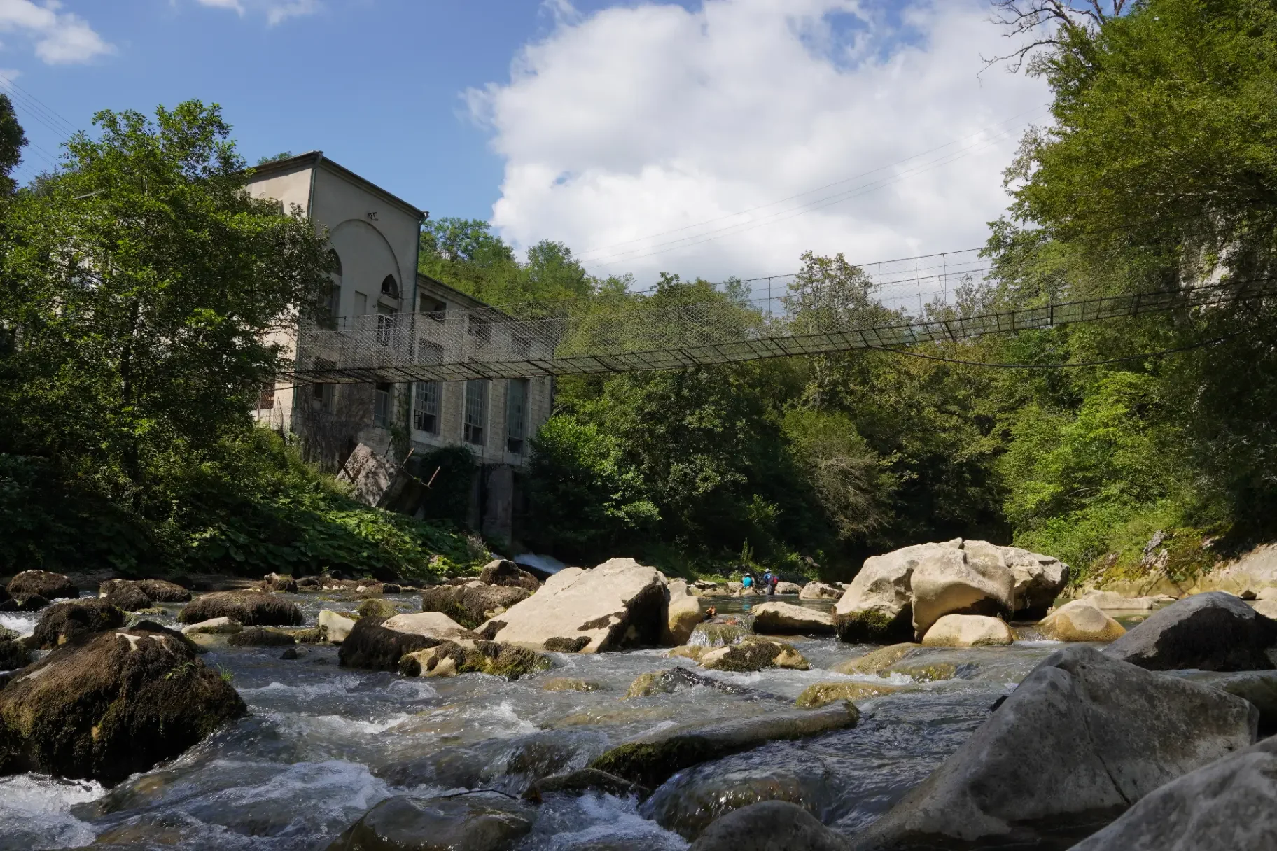 A river flowing over rocks with a suspension bridge above it, surrounded by green trees and a building in the background under a partly cloudy sky.