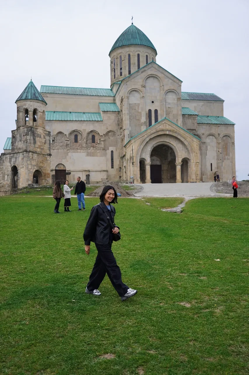 A woman in black walking on grass in front of a historic stone church with green rooftops, overcast sky, with other visitors nearby.