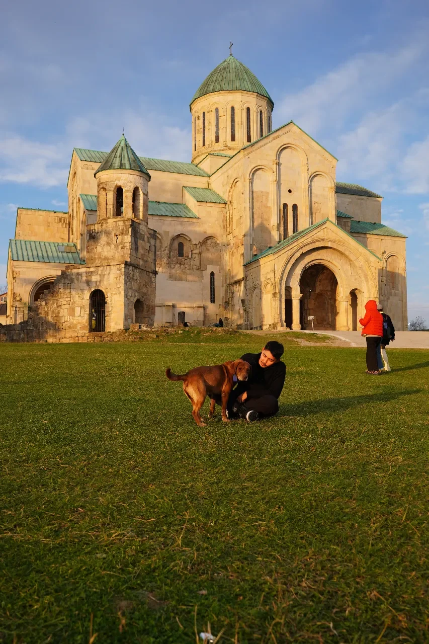 A person playing with a brown dog on a grass lawn in front of an ancient church with stone walls and multiple green roofs, under a partly cloudy sky.