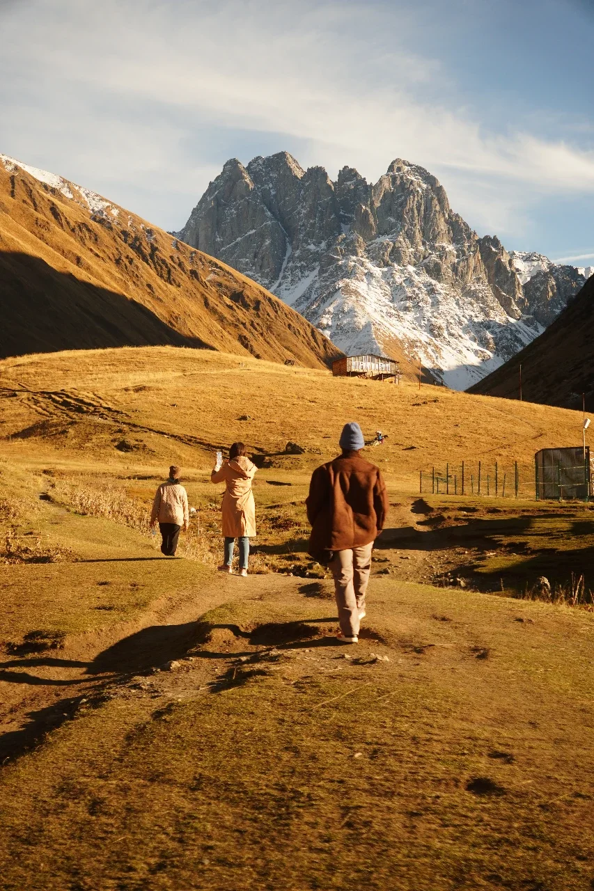 People walking on a dirt trail in a mountainous landscape with snow-capped peaks in the background, under a partly cloudy sky.
