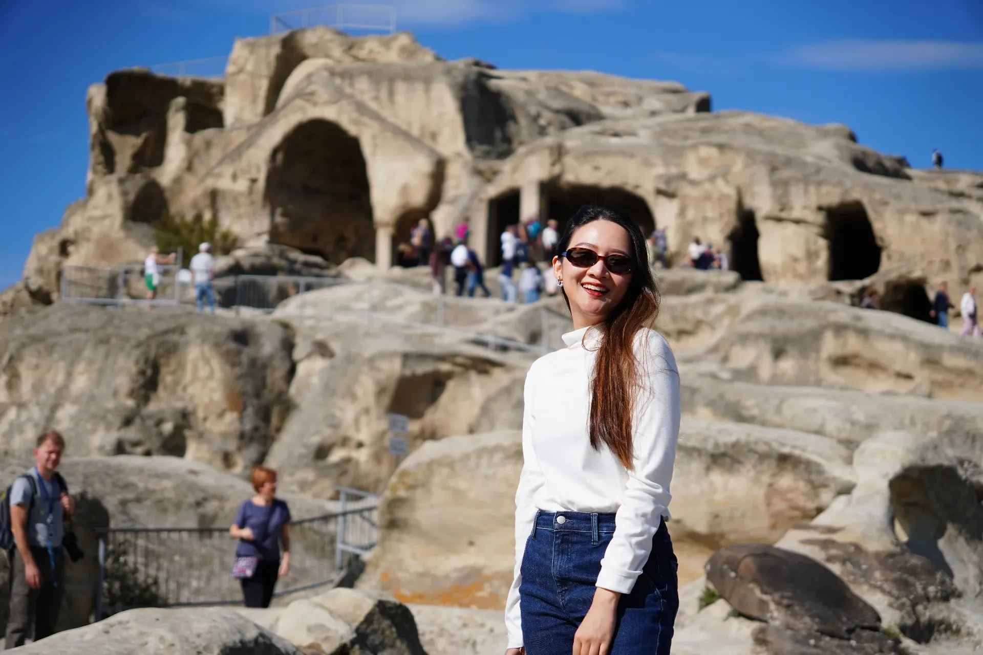 Smiling woman with long brown hair and sunglasses standing in front of an ancient rocky archaeological site with visitors in the background.