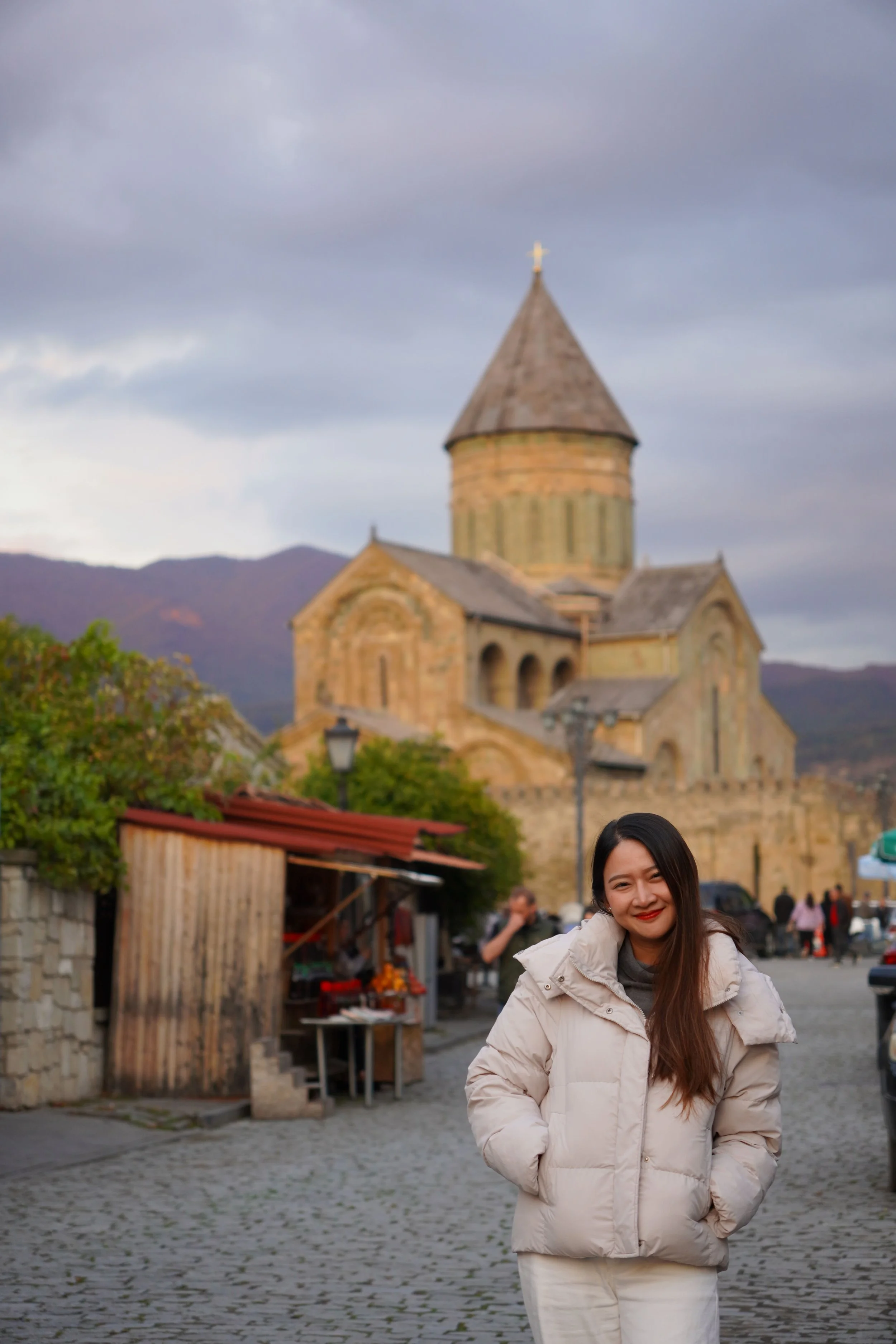 A woman in a white winter coat standing on a cobblestone street in front of a historic stone church with a rounded tower and cross on top, with mountains in the background.