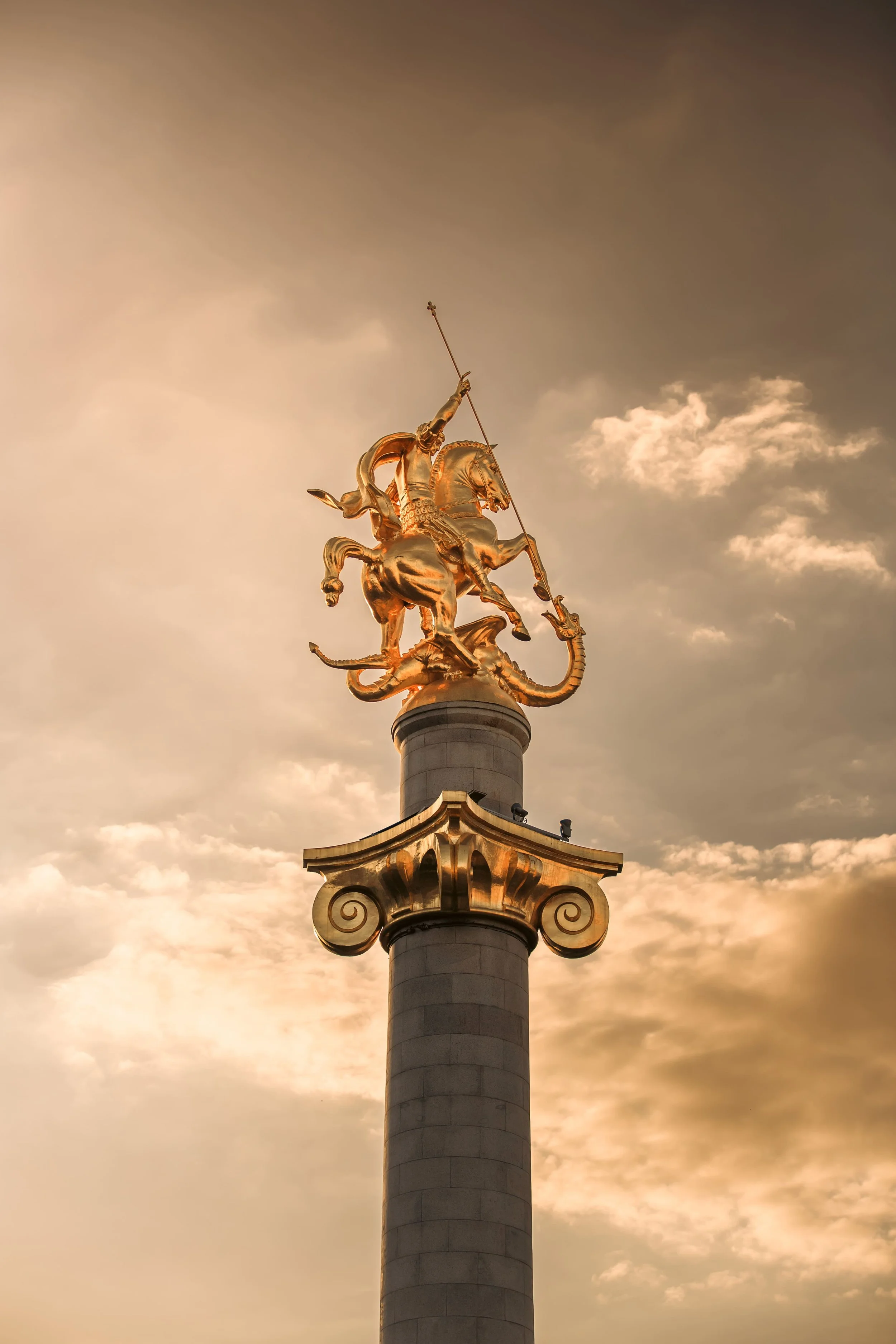 A golden statue of a warrior on a horse, holding a spear, atop a tall column against a cloudy sky.