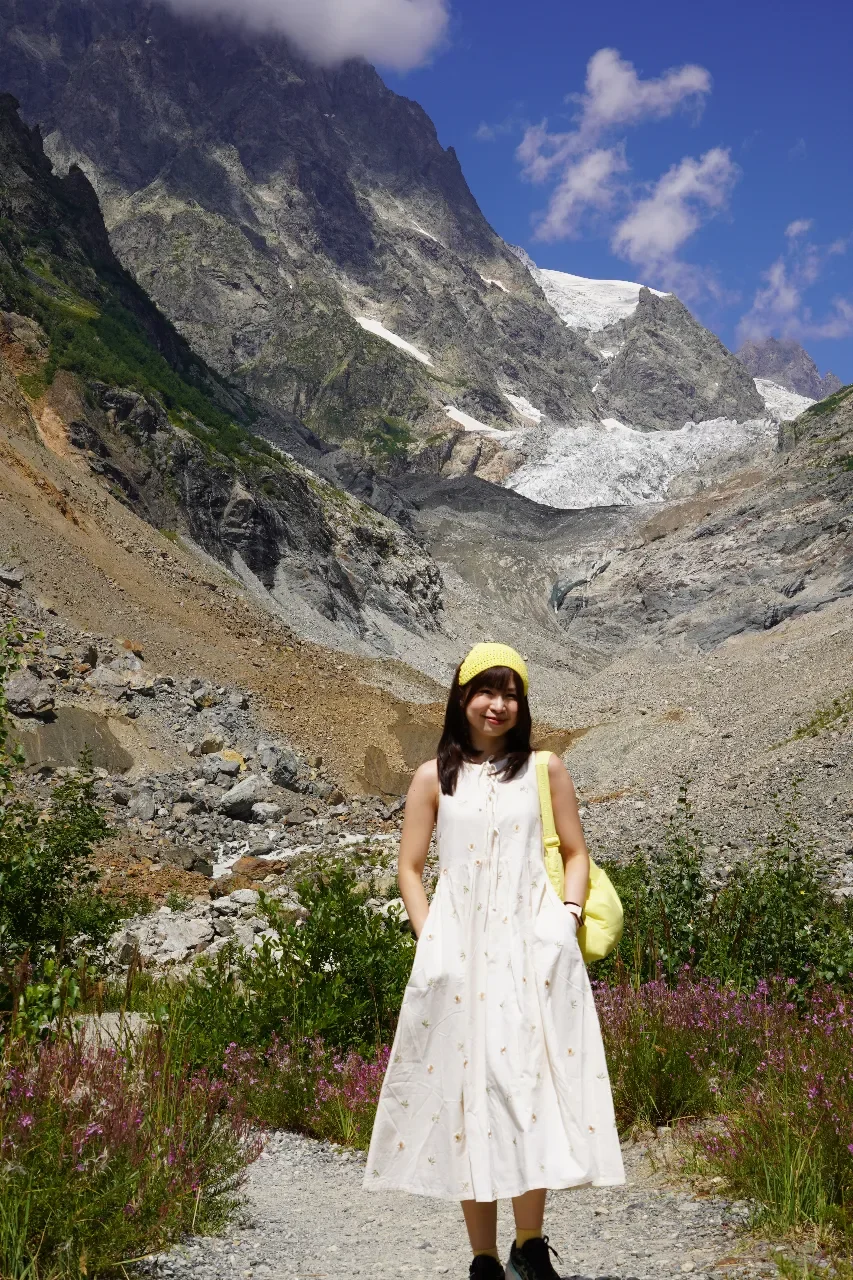 A woman standing on a trail at the base of a mountain with snow and glaciers. She is wearing a white dress, yellow hat, and carrying a yellow bag, with greenery and wildflowers around her.