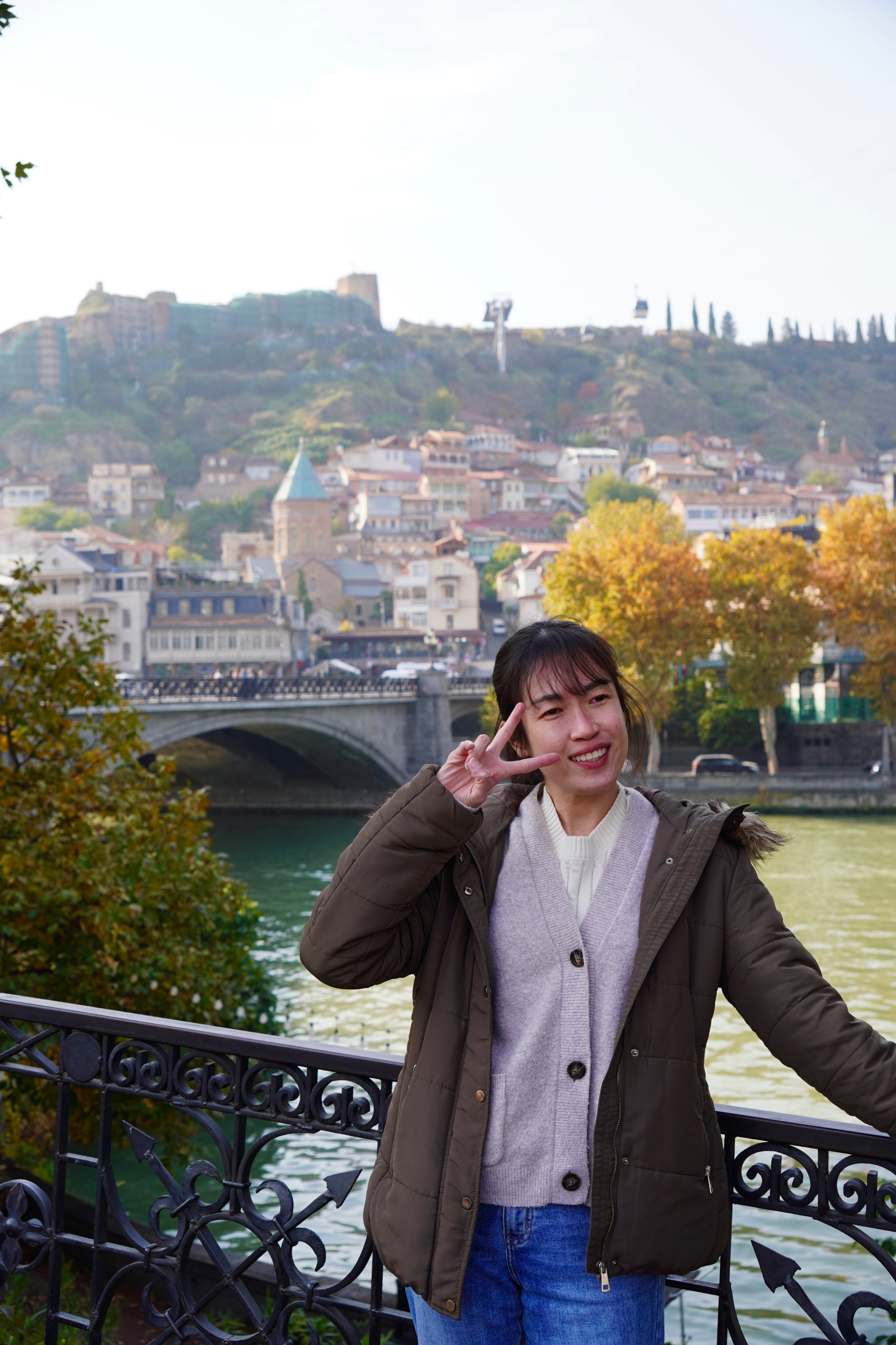 A woman making a peace sign near a river with a cityscape and hillside in the background.