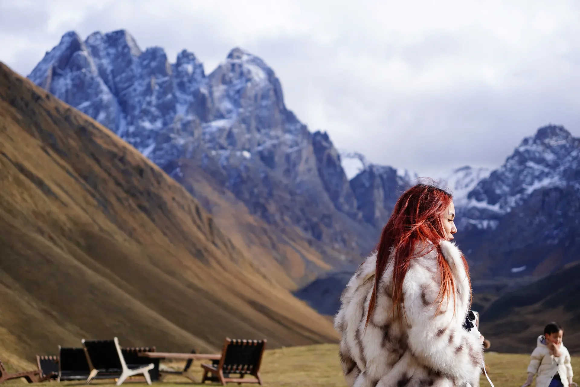A woman with red hair wearing a fur coat outdoors in a mountain landscape with snow-capped peaks and grassy slopes, and a young girl in the background.