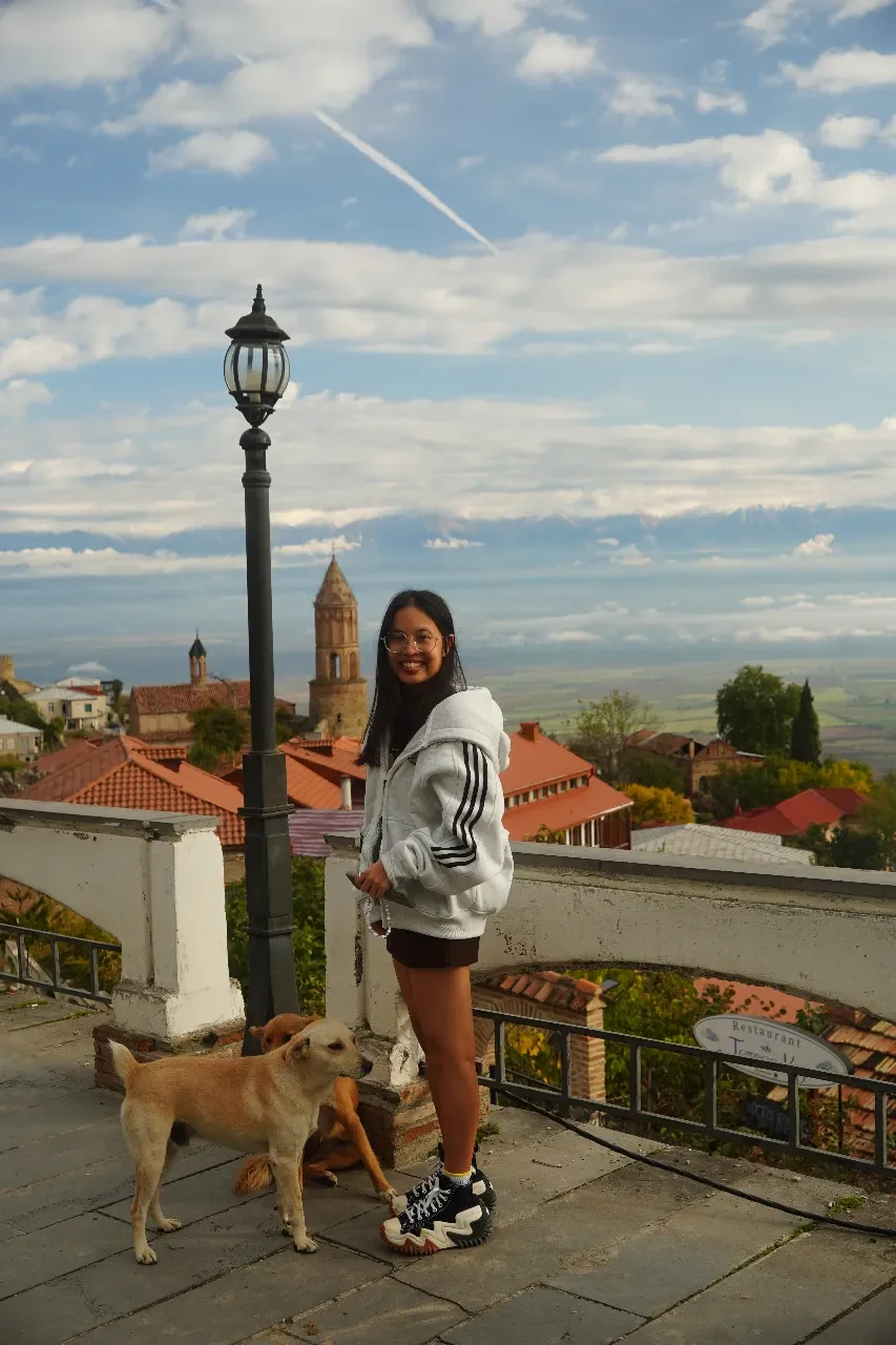 A smiling woman wearing a white hoodie with black stripes, shorts, and chunky sneakers standing outdoors on a terrace with two dogs, overlooking a town with red-tiled roofs, a church tower, and a scenic landscape under a partly cloudy sky with a cont