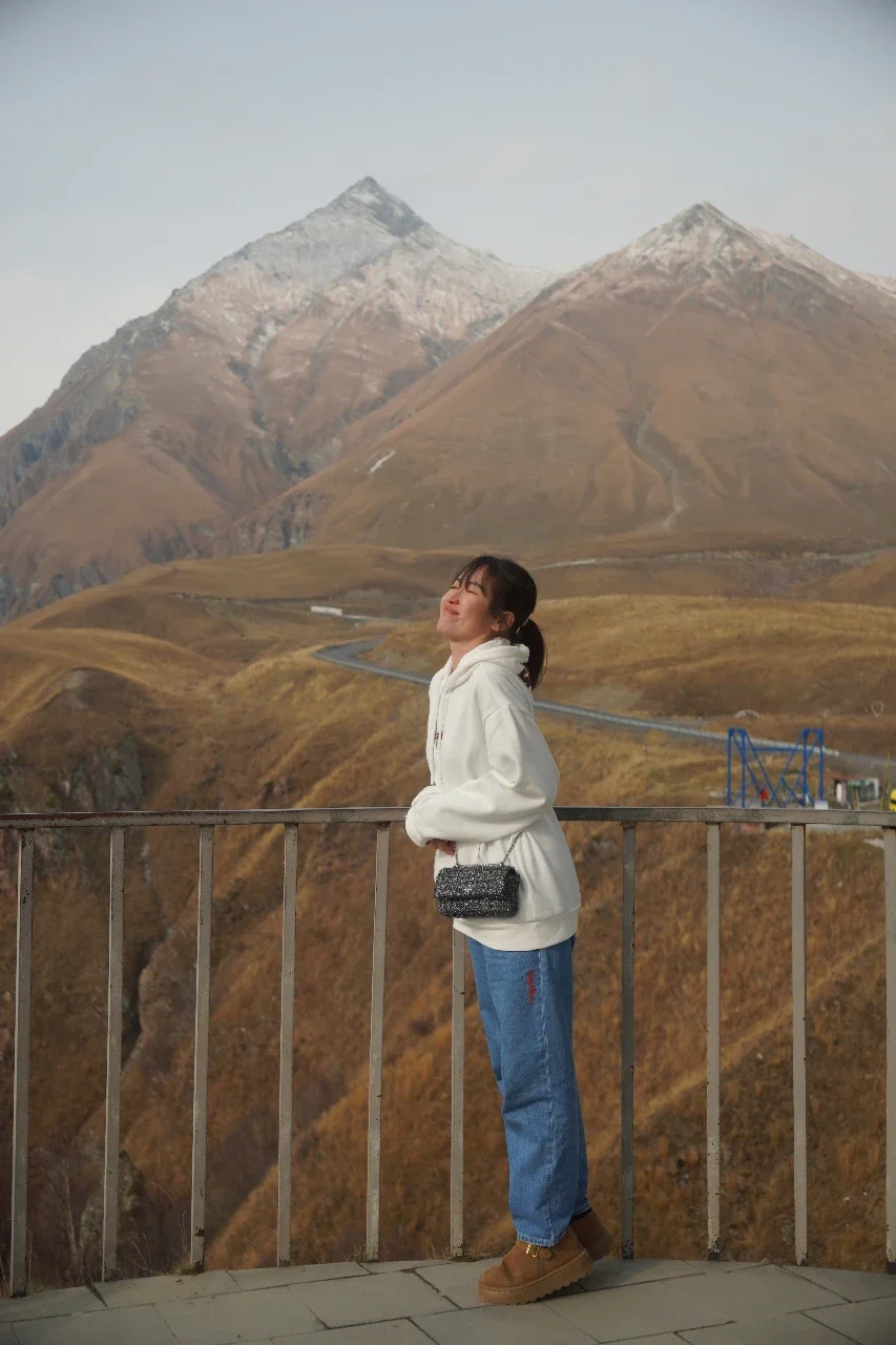 Young woman in a white hoodie and jeans standing on a balcony with mountain peaks in the background.
