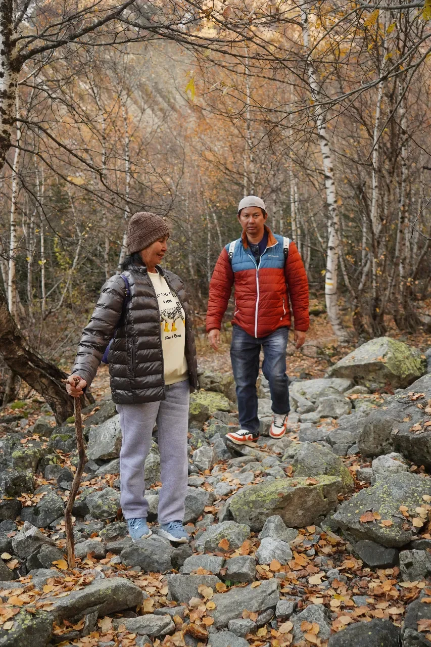 Two people hiking on a rocky trail in a forest during autumn, surrounded by trees with fall-colored leaves.