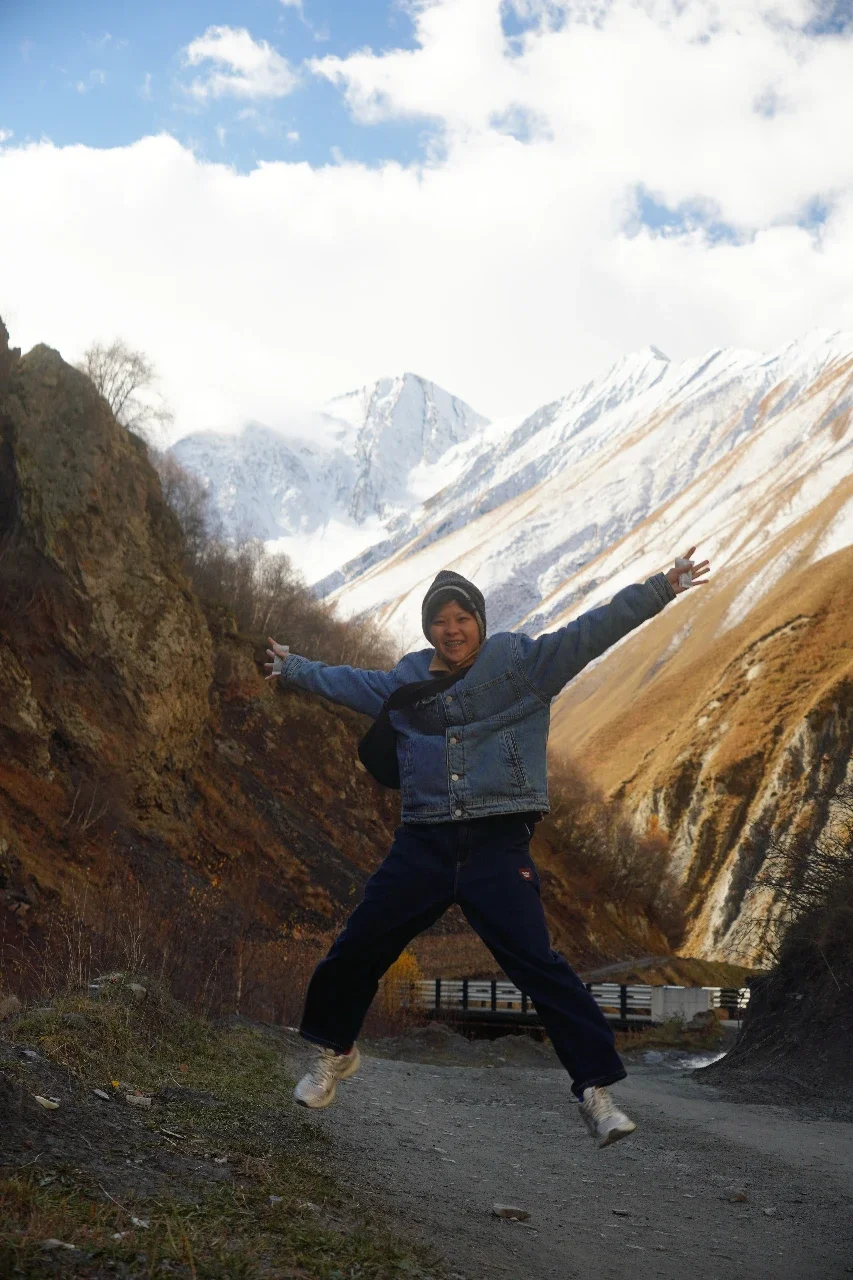 Child in a blue denim jacket and dark pants jumping with arms wide open in a mountainous landscape with snow-covered peaks and a partly cloudy sky.