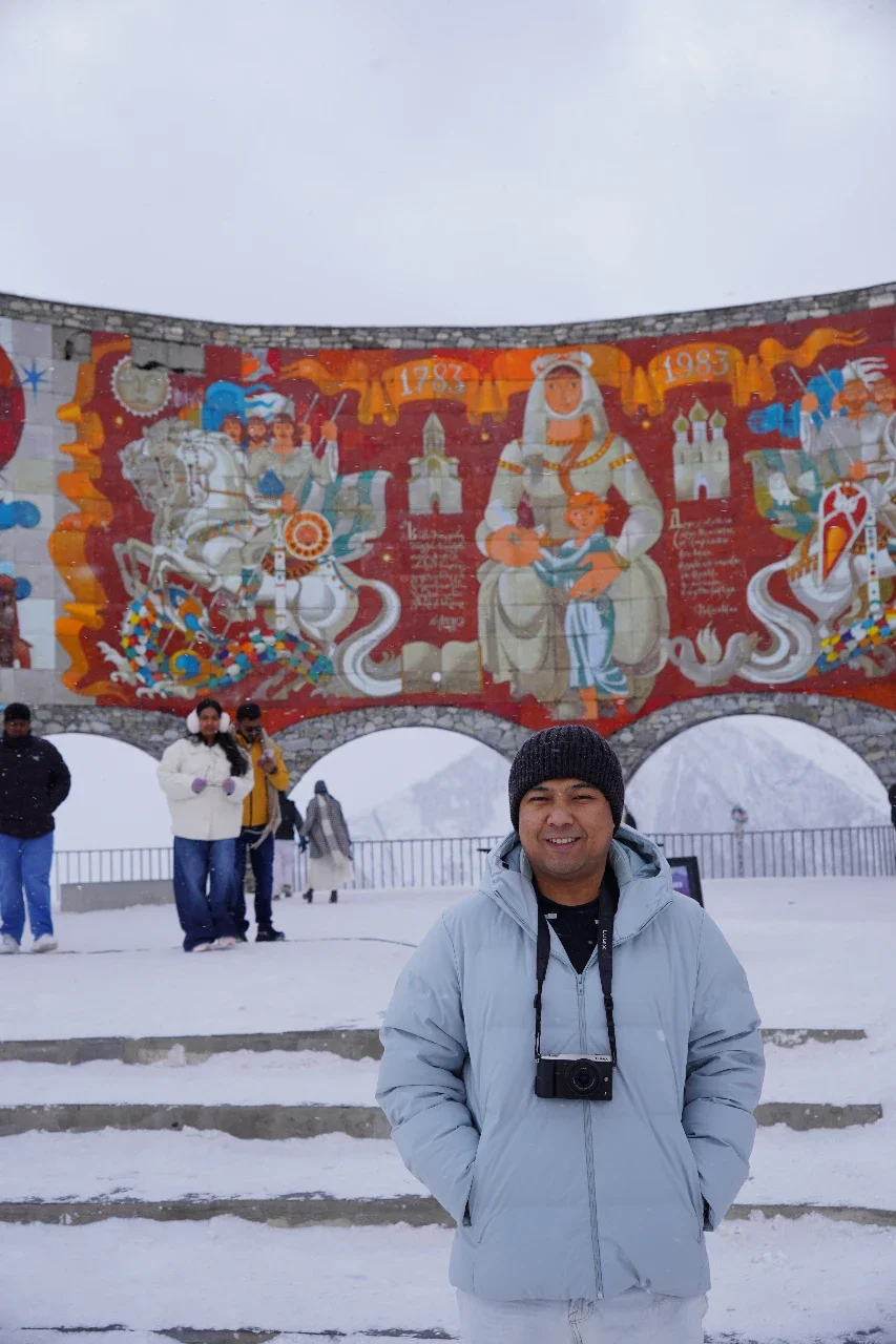 A man in a gray winter jacket and black beanie smiling in front of a colorful mural with historical figures, while others stand on snowy steps behind him.