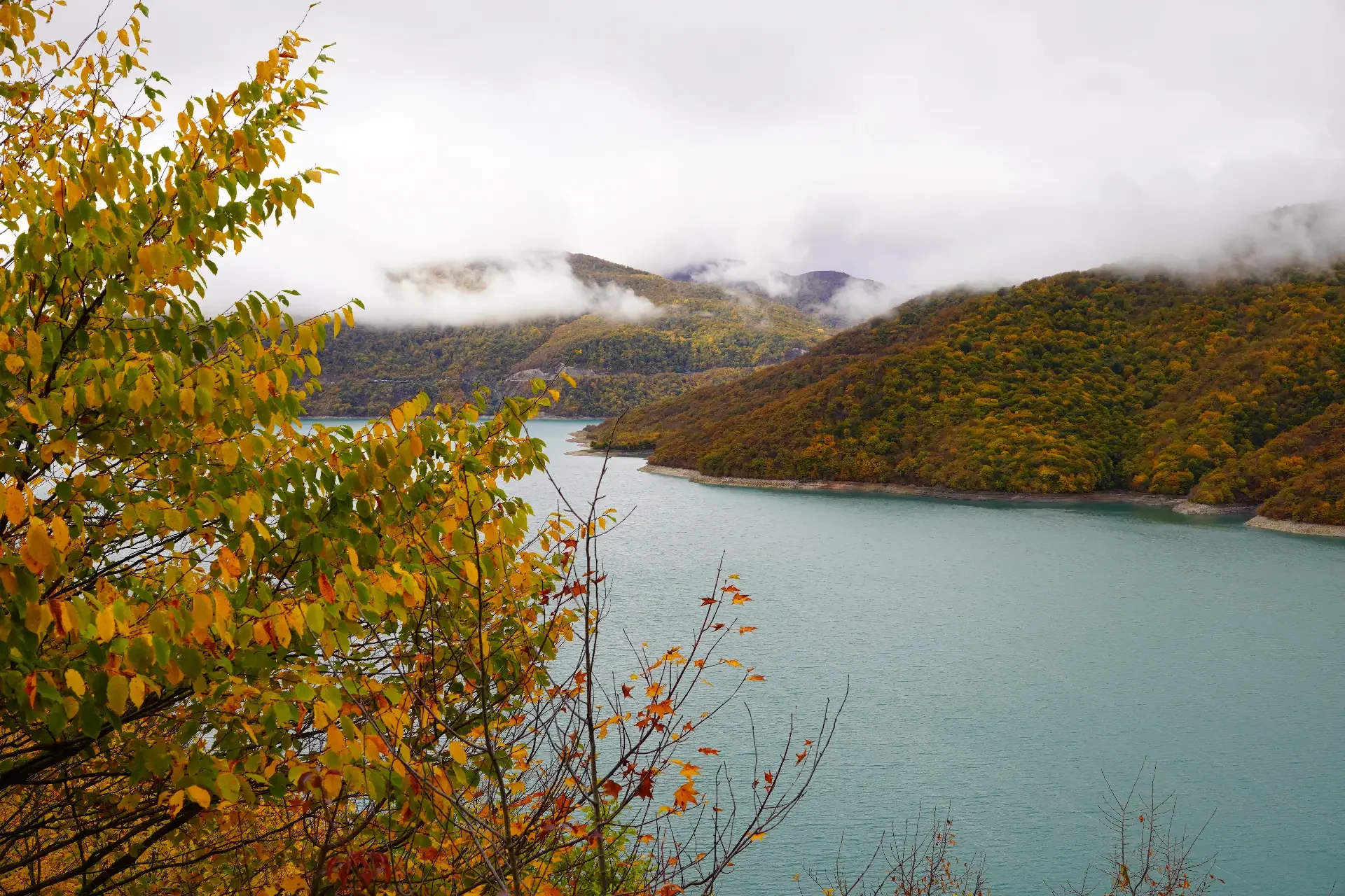 Scenic view of a lake surrounded by forested hills with fall foliage, cloudy sky, and mist over the mountains.