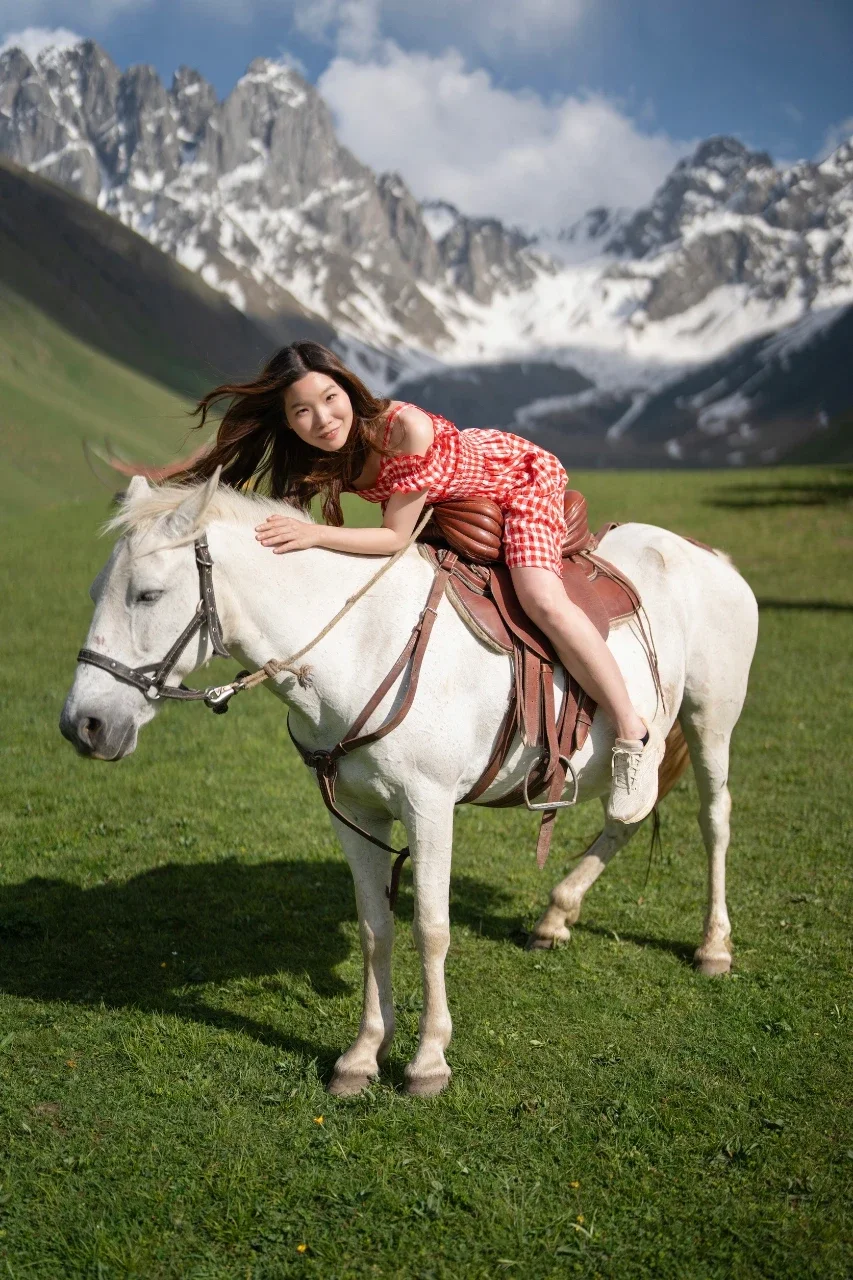 Young woman lying on a white horse in a green meadow with snow-capped mountains in the background.