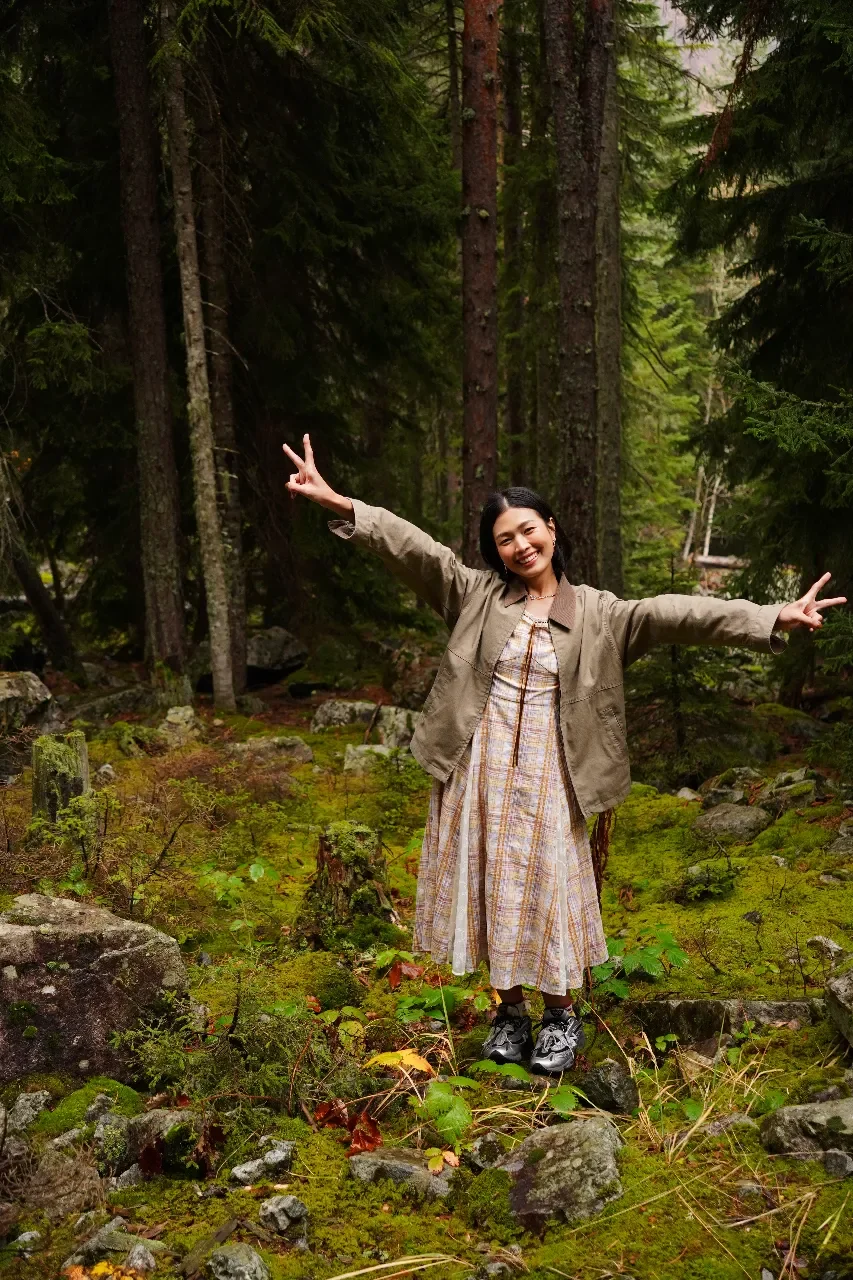 A woman standing on rocks in a lush green forest with tall trees, smiling and making peace signs with both hands.