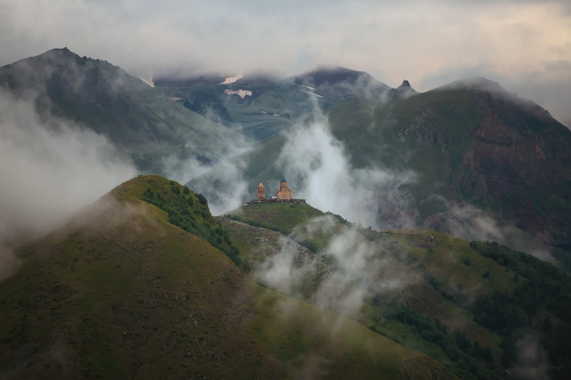Mountains with green slopes, fog, and a small church or castle on a hilltop.