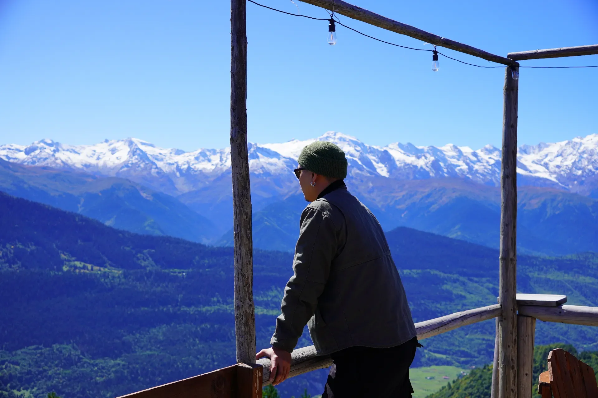 A person wearing a green beanie, sunglasses, and a dark jacket leaning on a wooden railing, looking at snow-capped mountains and lush green hills under a clear blue sky.