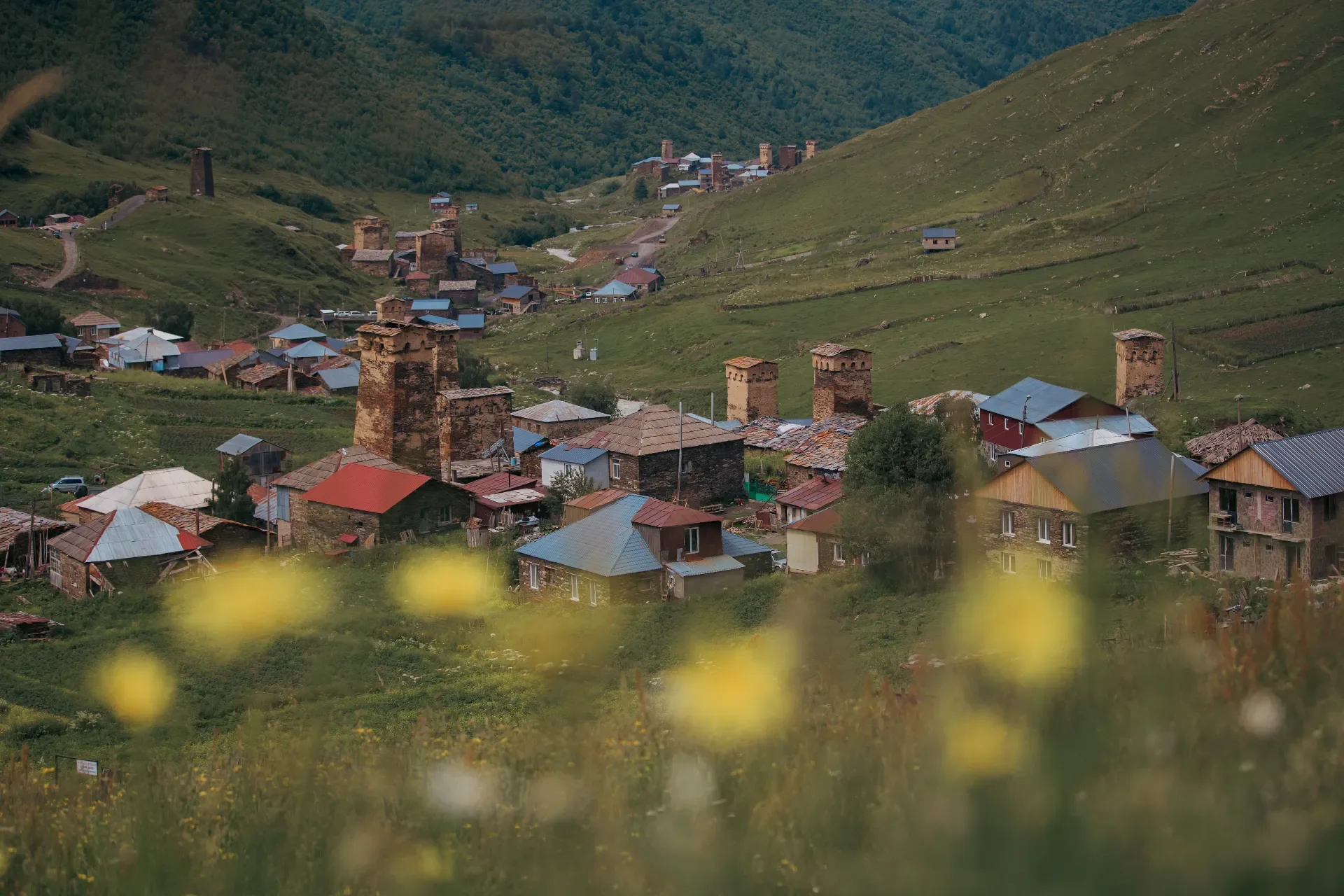 Scenic view of a small village with stone towers and houses situated in a lush green valley surrounded by hills and mountains.
