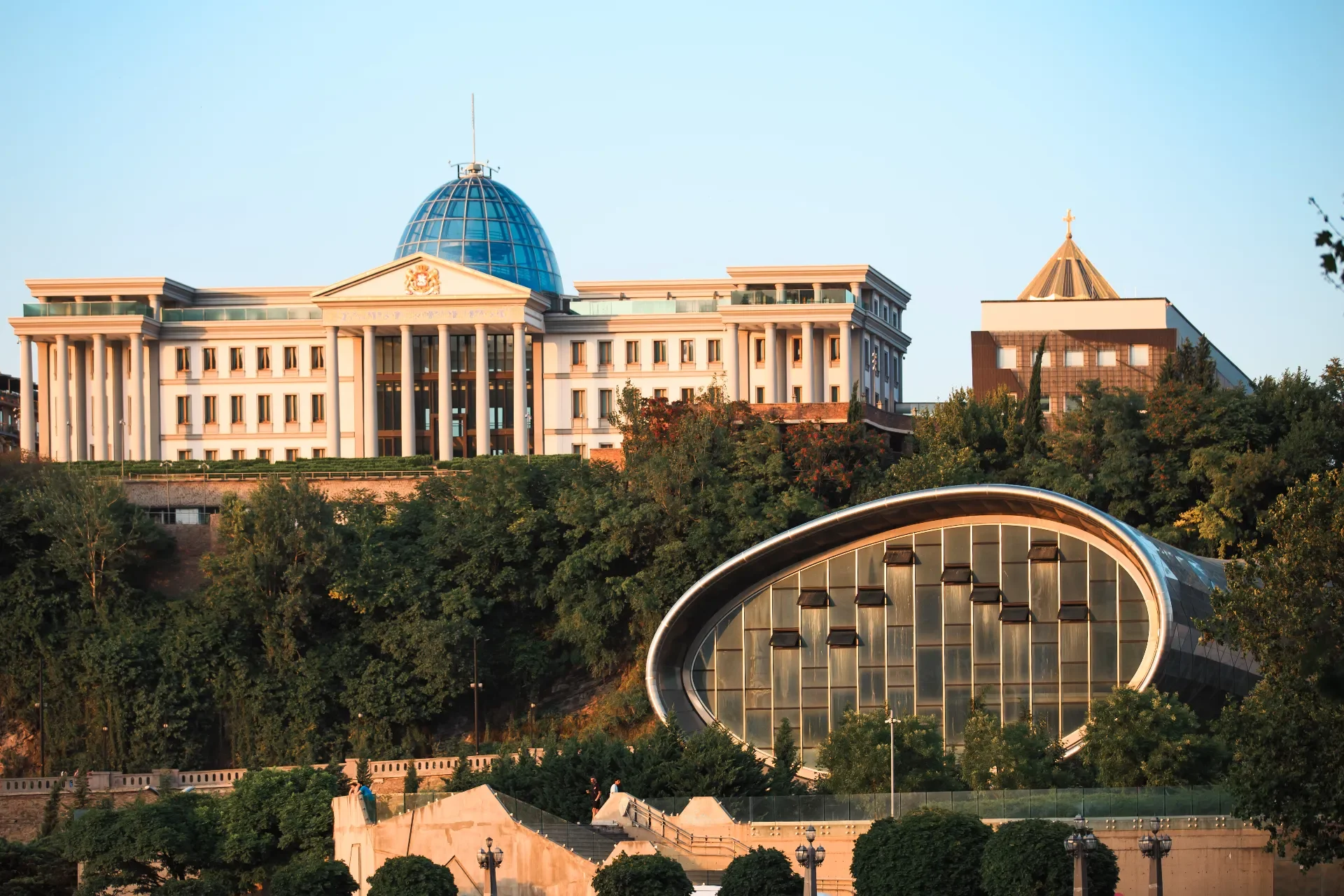 A large white building with columns and a blue dome on top, situated on a hillside with trees. There is also a modern, oval-shaped glass structure in the foreground.