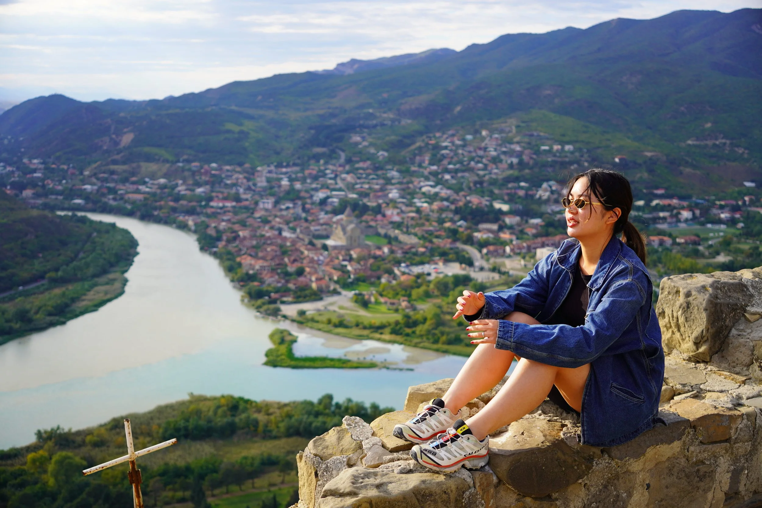 A woman sitting on a rocky ledge overlooking a river and a town surrounded by mountains.