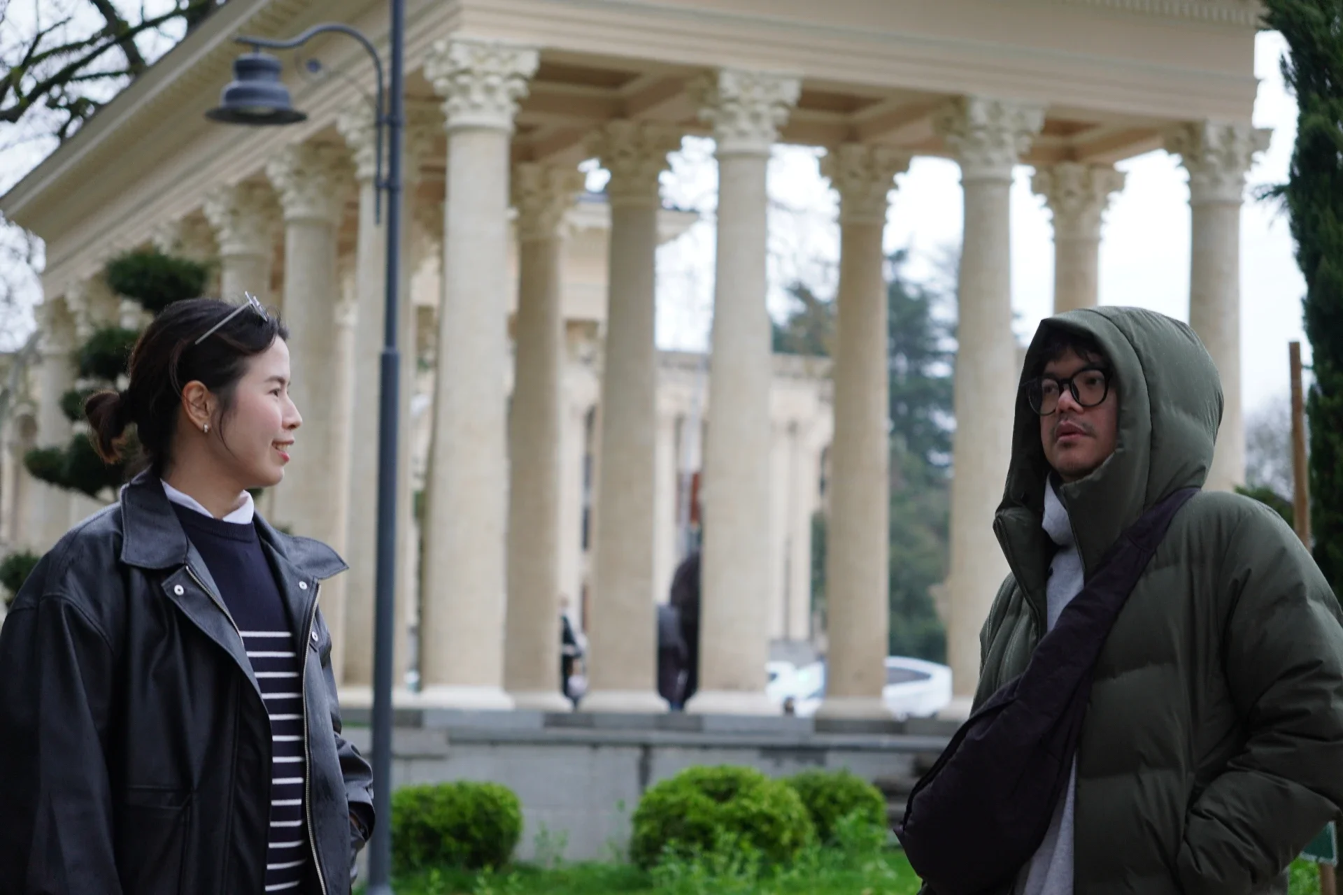 A woman and a man engaged in conversation outdoors in front of a building with classical columns. The woman has dark hair tied back, wearing a dark jacket and striped shirt. The man is wearing glasses and a green hooded jacket. There are small bushes