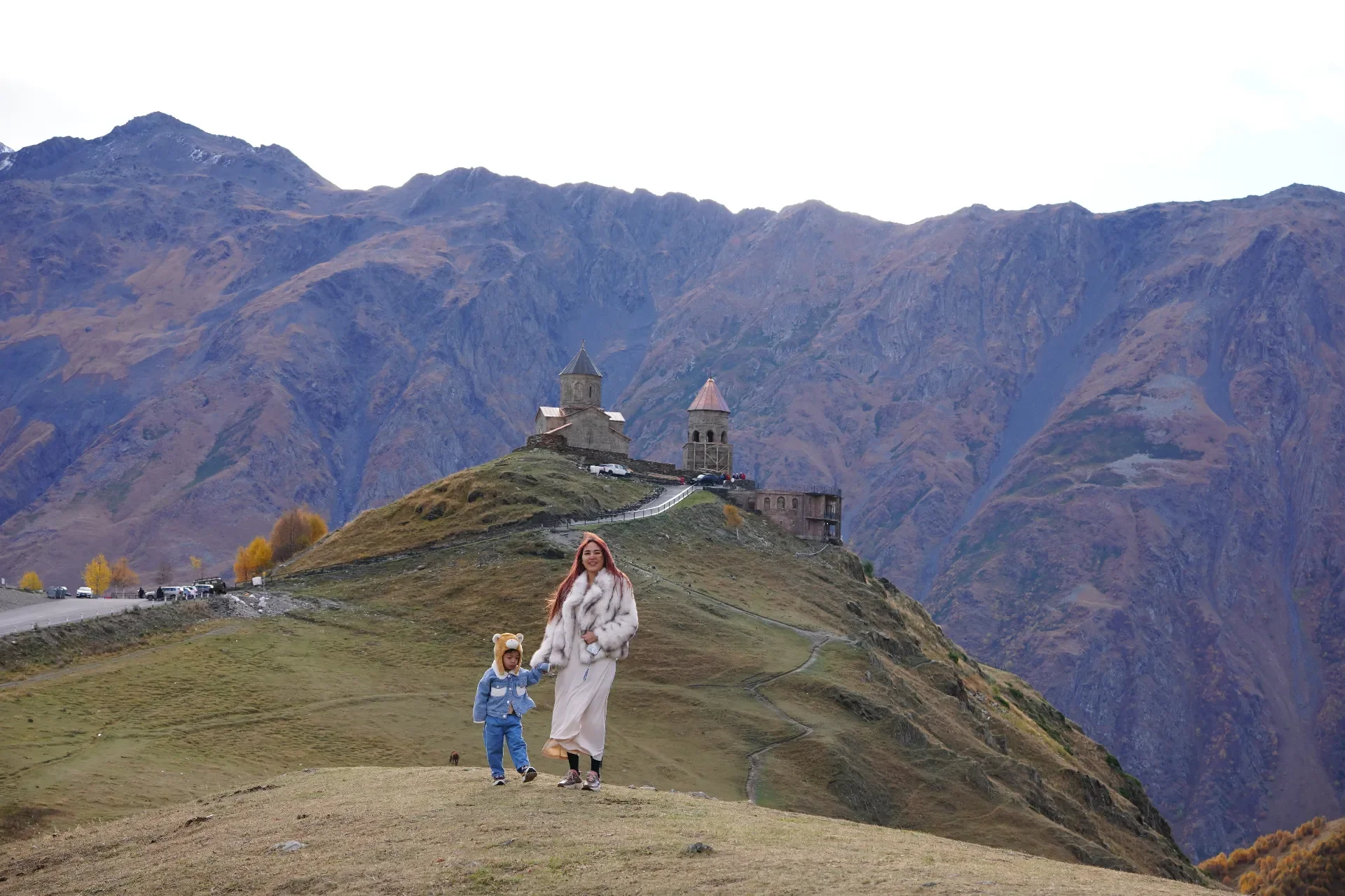 A woman and a child walking hand in hand on a grassy hill with a stone church on a hilltop and mountains in the background.