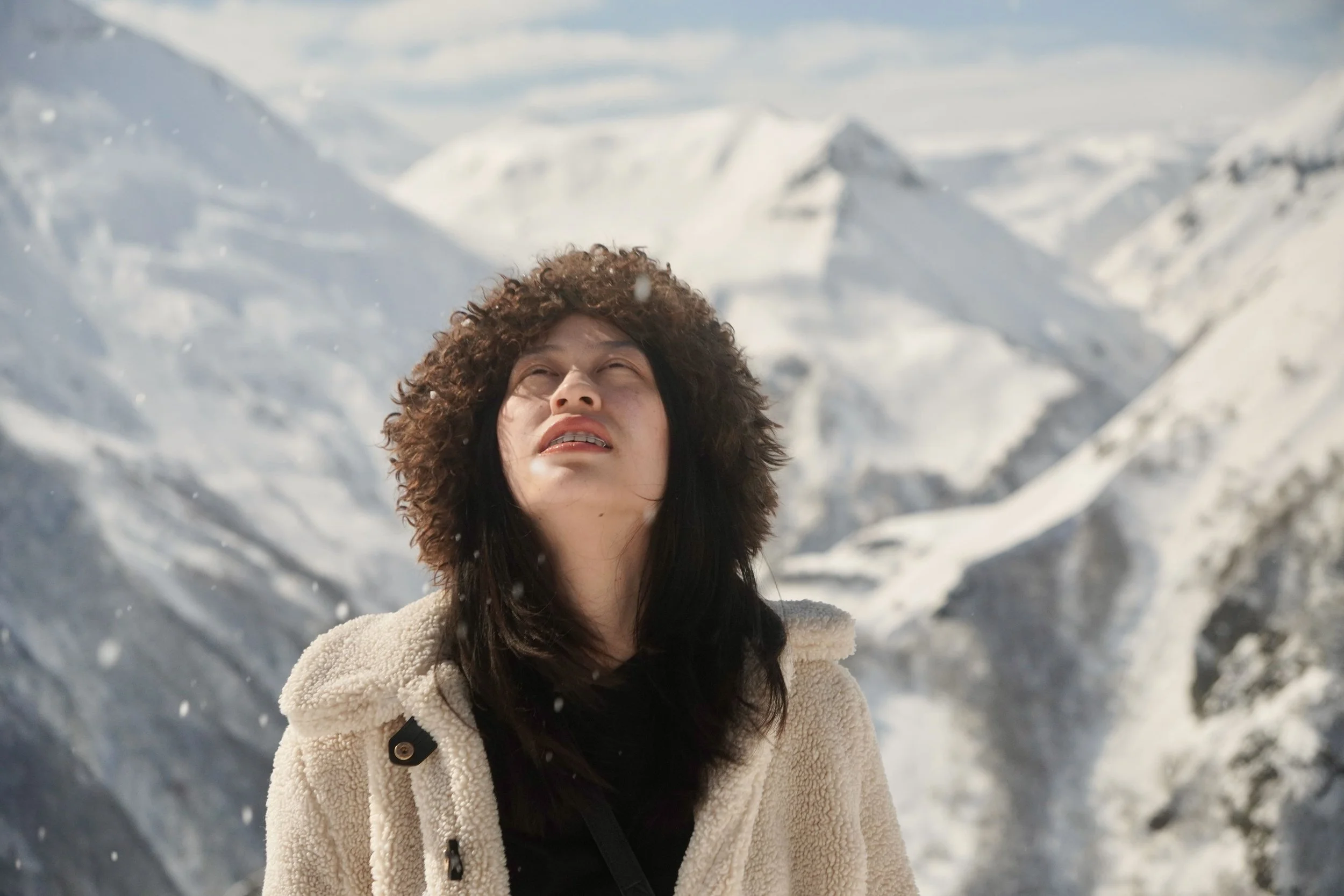 Woman with curly hair and a beige fleece jacket stands outdoors in a snowy mountain landscape, looking up with a concerned expression.
