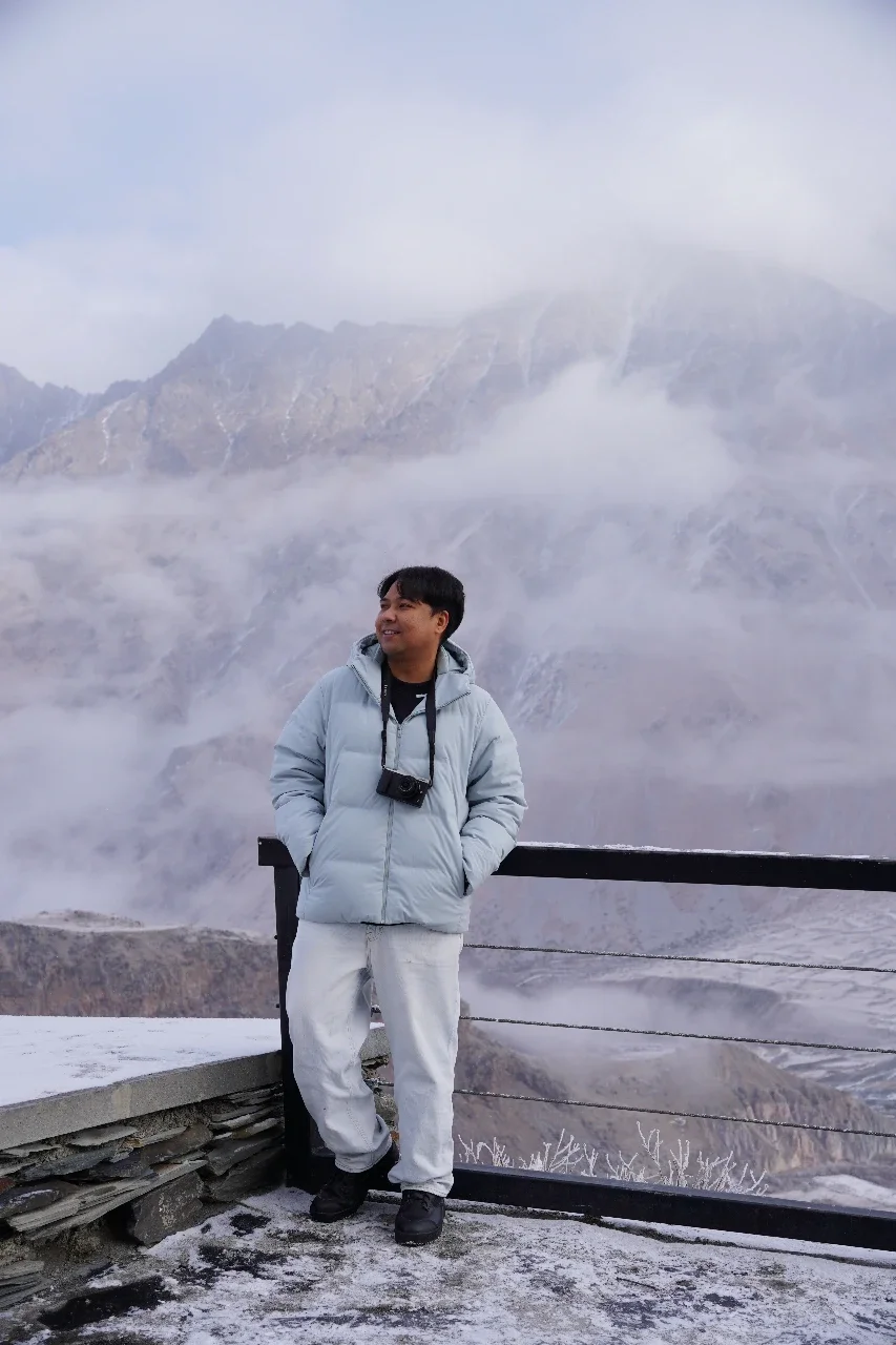 A man standing on an outdoor viewpoint in a snowy, mountainous landscape with fog and rugged peaks in the background, wearing a light gray winter jacket and white pants, with a camera hanging around his neck.