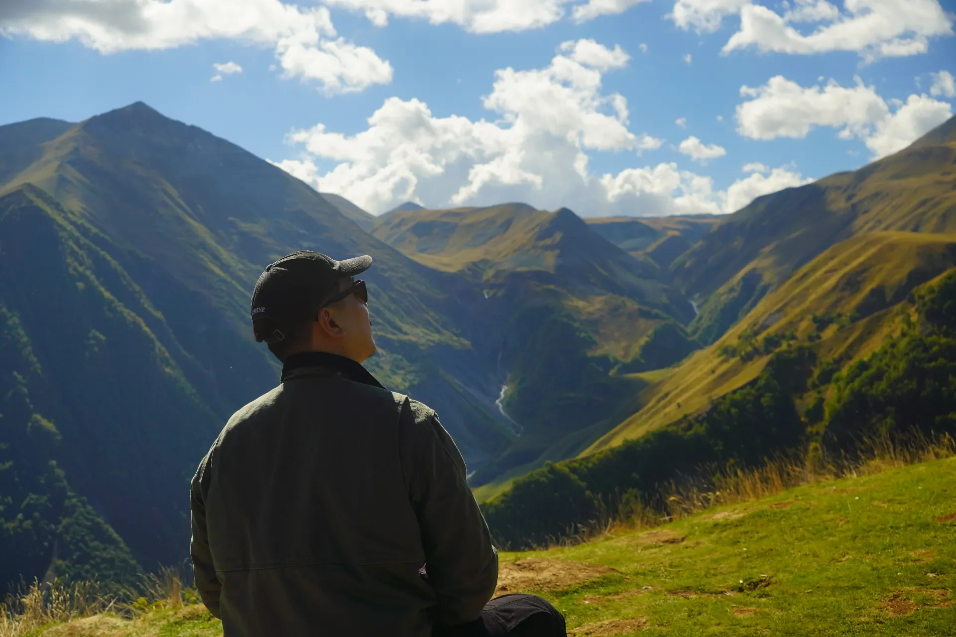 A person wearing a dark jacket and cap sitting on a grassy hill, overlooking a mountainous landscape with green valleys and a waterfall, under a partly cloudy sky.