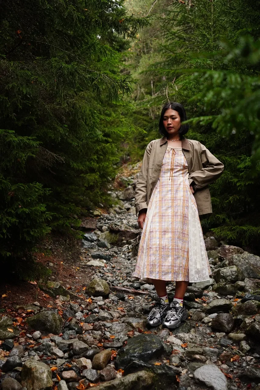 A woman standing on a rocky forest trail surrounded by dense green trees, wearing a dress, jacket, and sneakers.