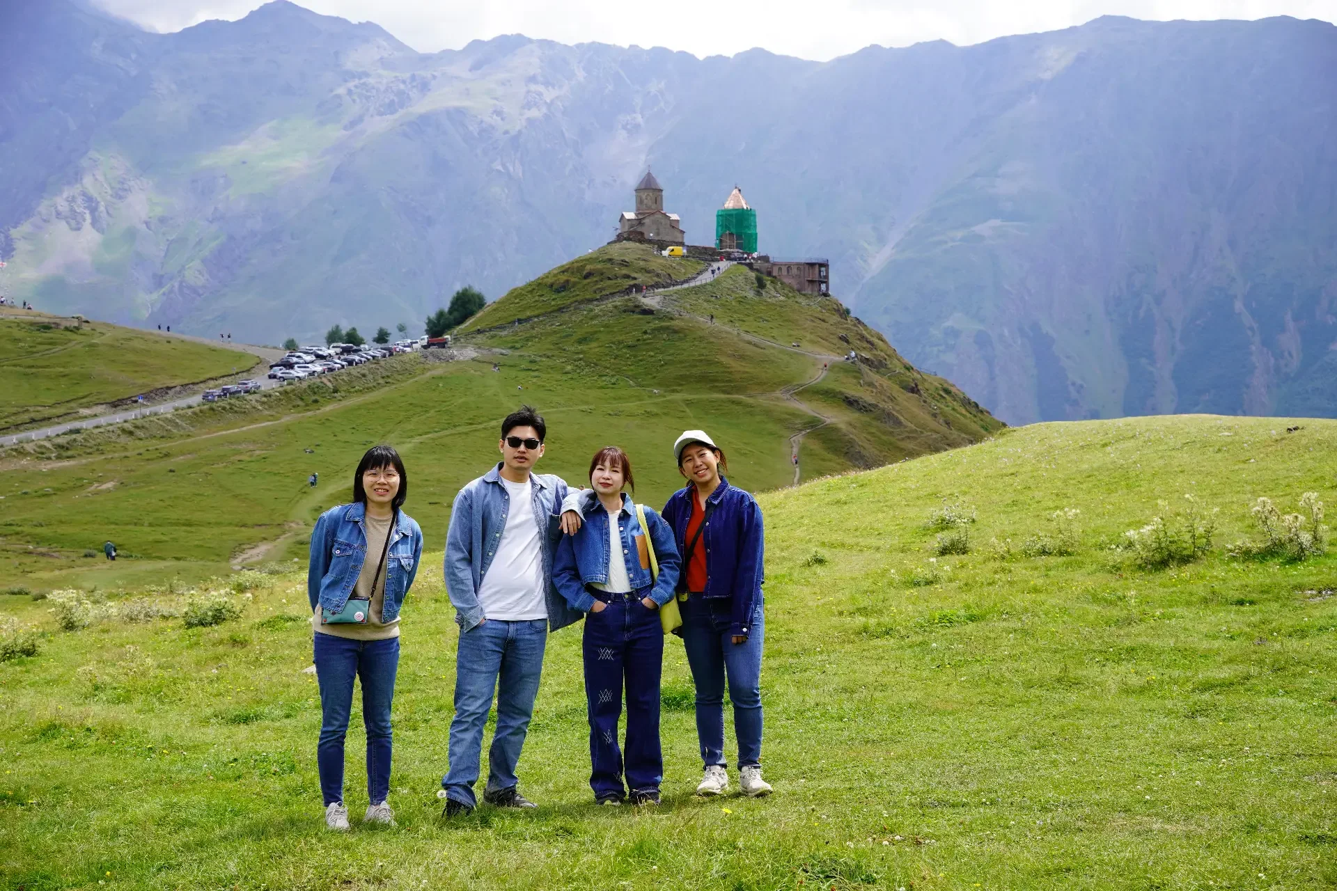 Four people standing on green grass with mountains and a church in the background.