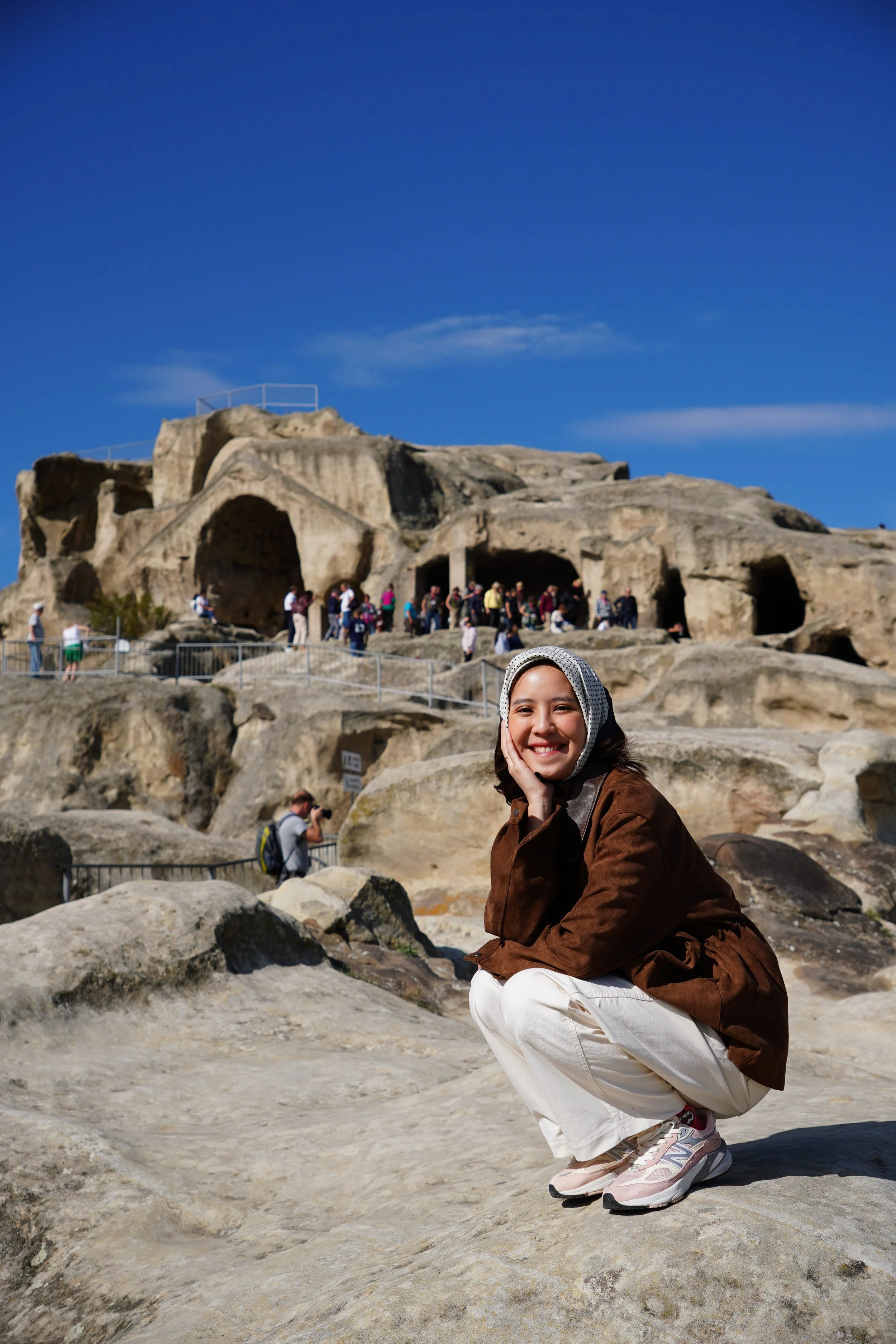 A smiling woman squatting on rocks in front of ancient cave dwellings and a crowd of tourists at a historical site under a clear blue sky.