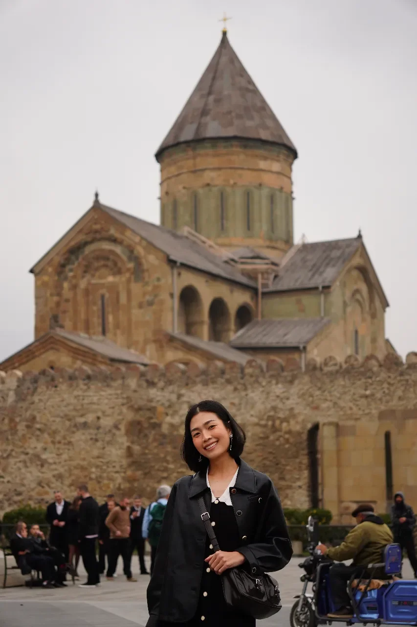 A young woman smiling in front of an old stone church with a large conical roof and a cross on top, with a group of people and a man on a motorized wheelchair in the background.