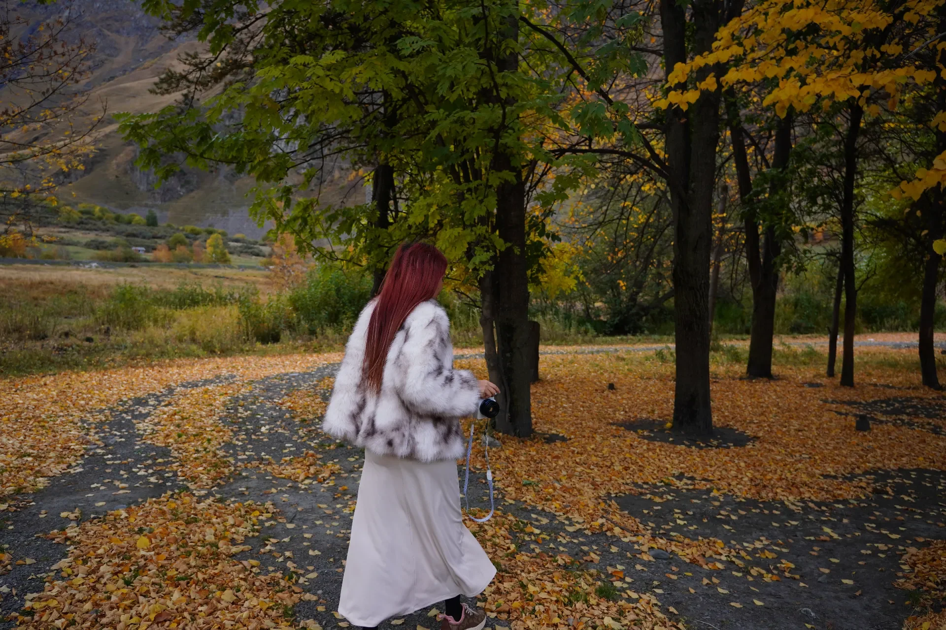 A woman with long red hair wearing a white fur coat and a white skirt walking through a fall landscape with yellow leaves on the ground and trees, holding a camera.