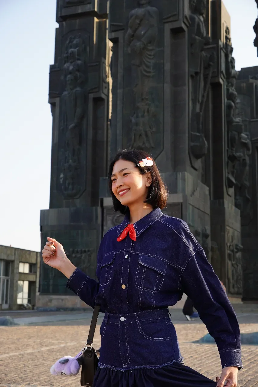Smiling woman with short black hair wearing a denim outfit, a red necktie, and Hello Kitty hair clips, standing outdoors in front of a large dark monument with detailed carvings.