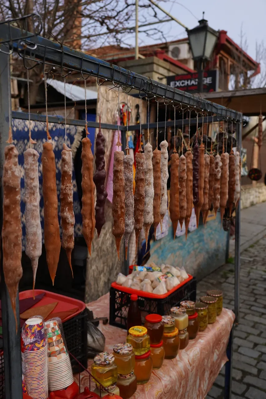 Dried, stick-shaped foods hanging on a metal rack at an outdoor market, with jars of honey and other products on a table below.