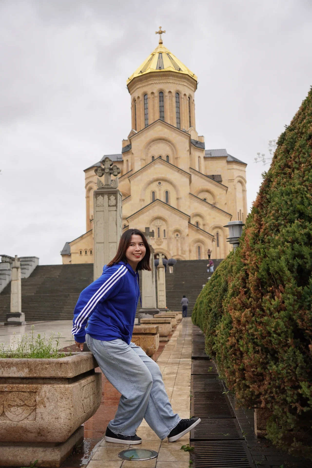 Young woman in a blue hoodie and light gray sweatpants posing on a stone ledge in front of a church with a golden dome, cloudy sky, and well-maintained bushes.
