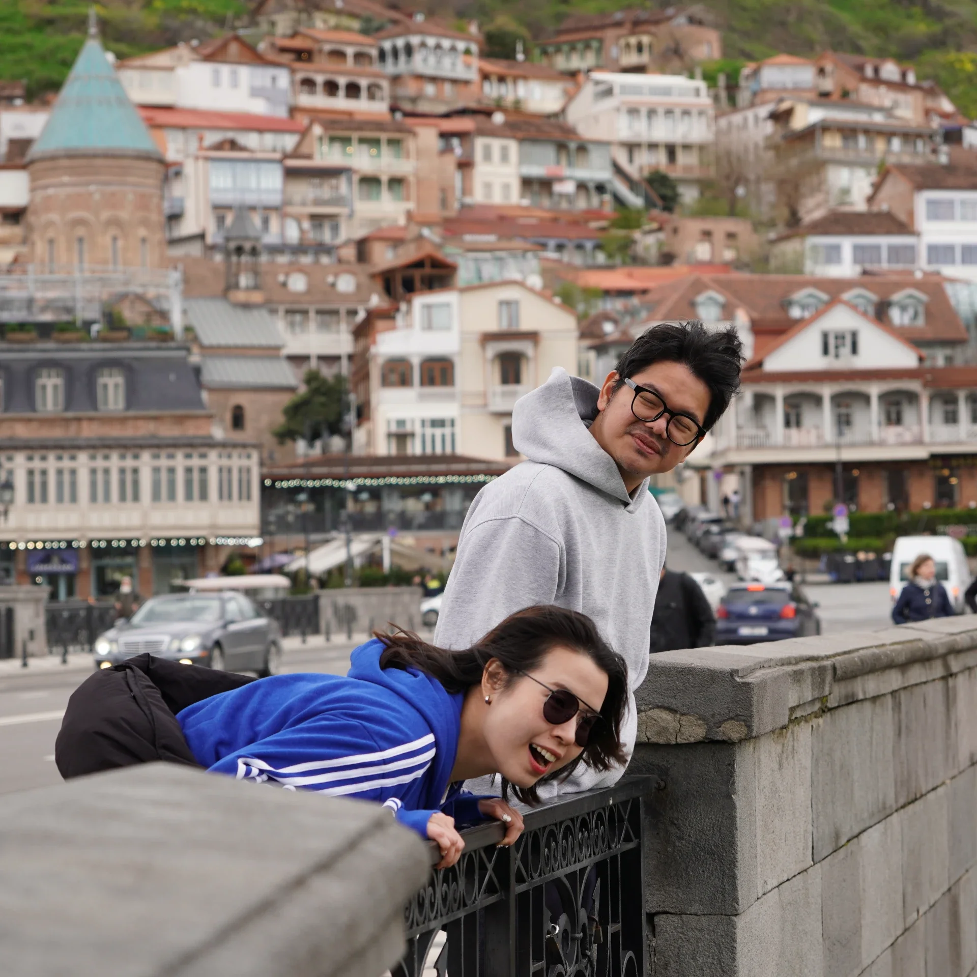 Two people leaning over a stone and iron fence, laughing and smiling, with a hillside filled with houses in the background.