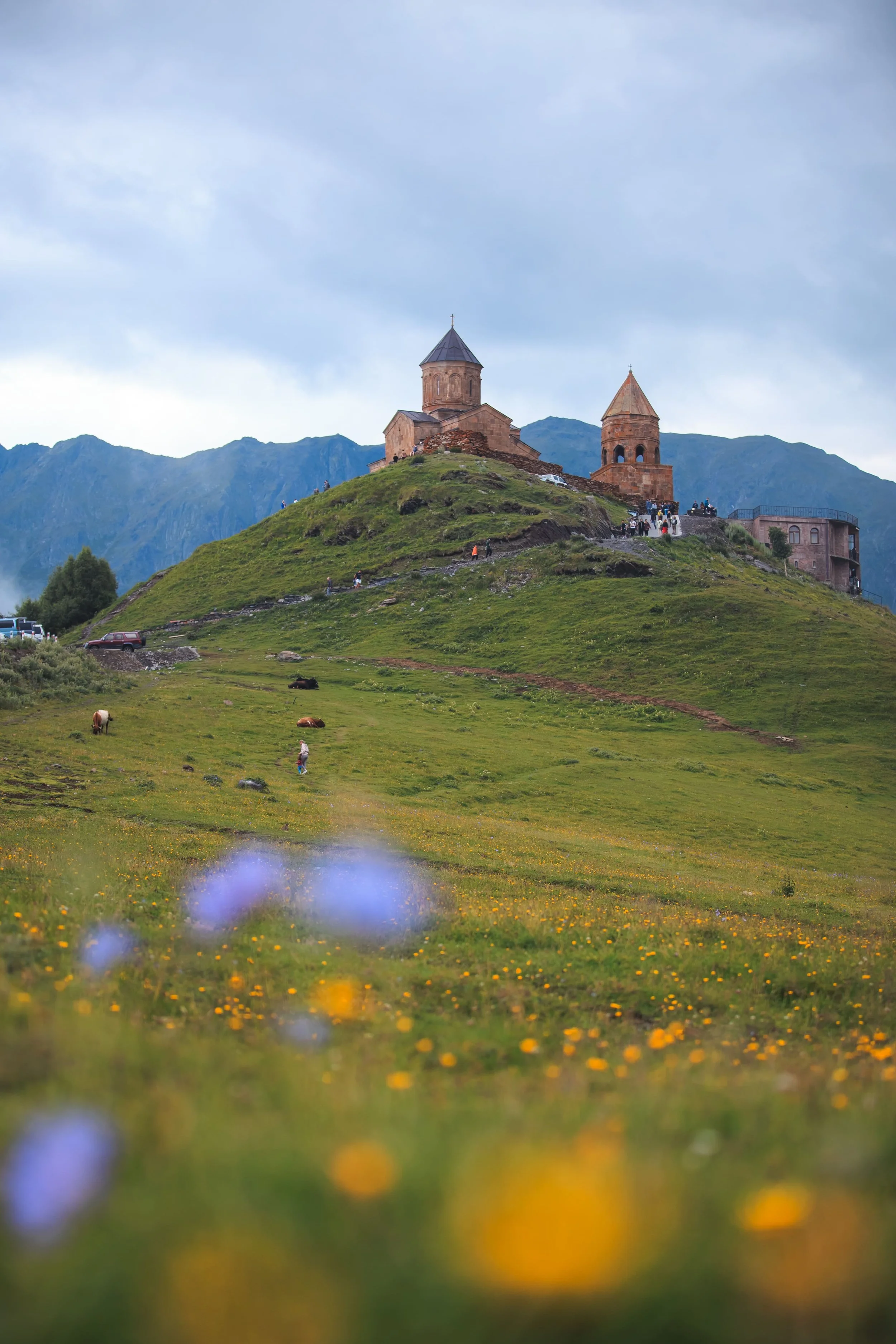 A church situated on a hilltop surrounded by mountains, with a green grassy field in the foreground dotted with yellow and purple flowers and a few people walking.