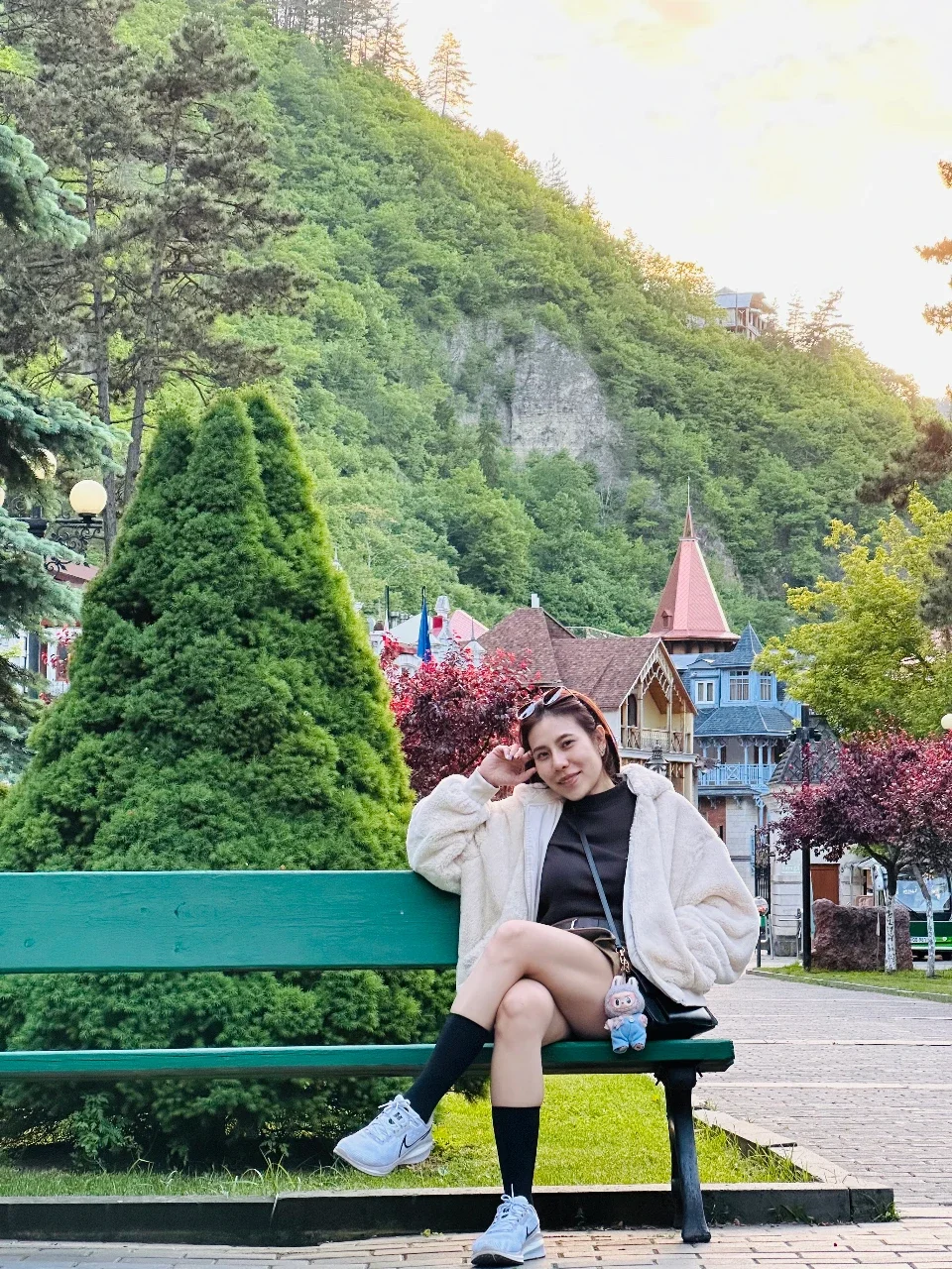 Young woman sitting on a park bench with lush greenery, colorful houses, and a hillside in the background.