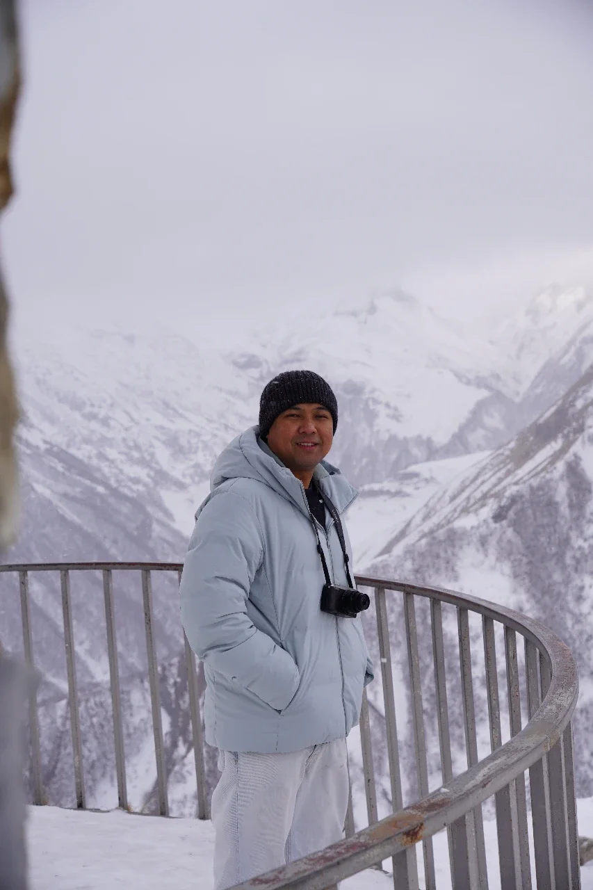 Man in winter clothing standing on a snowy mountain overlook with snow-covered peaks in the background.