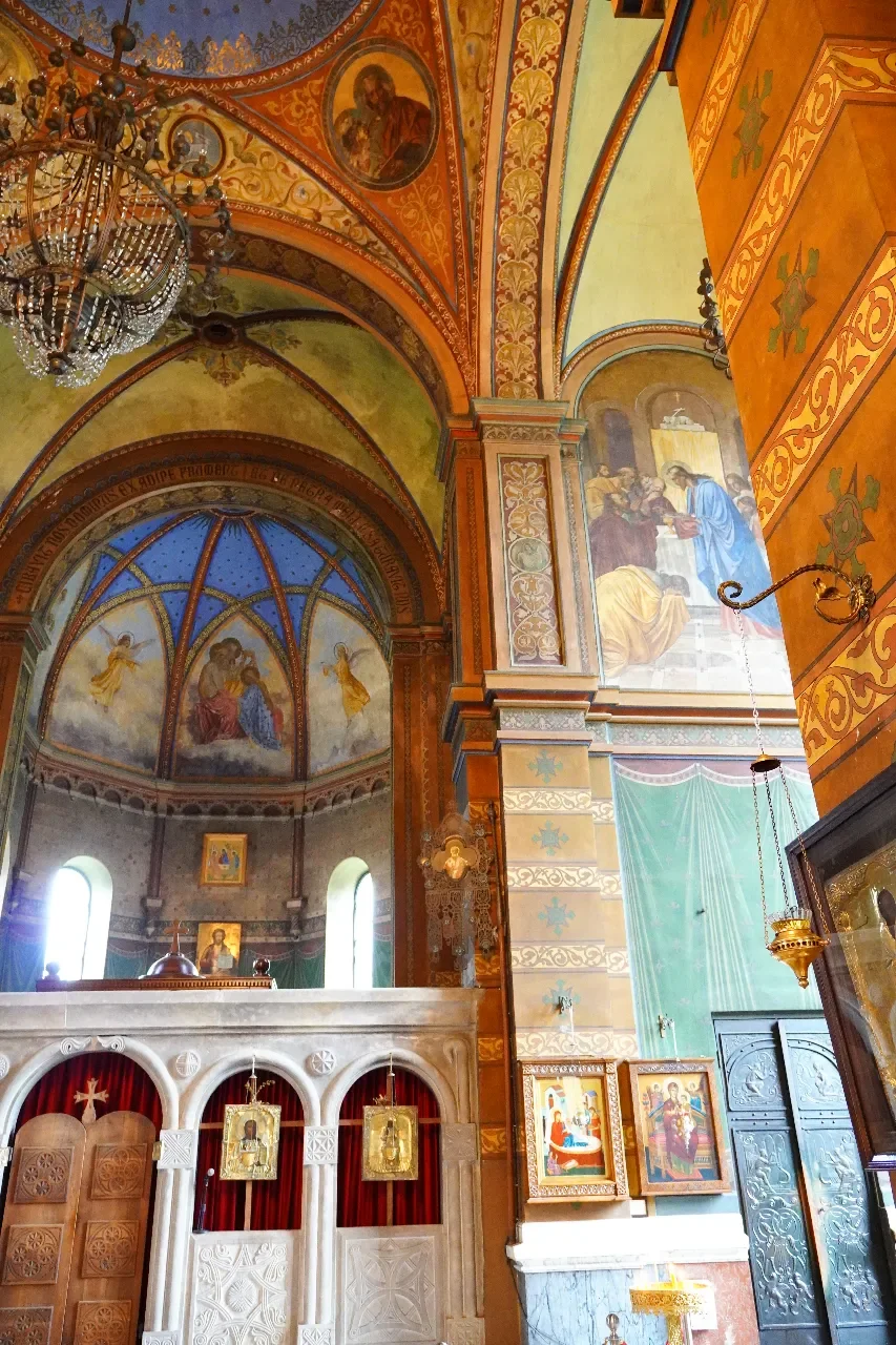 Inside a richly decorated Orthodox church, featuring ornate ceiling murals with angels and religious figures, a large chandelier, painted icons, and an altar area with religious icons and curtains.