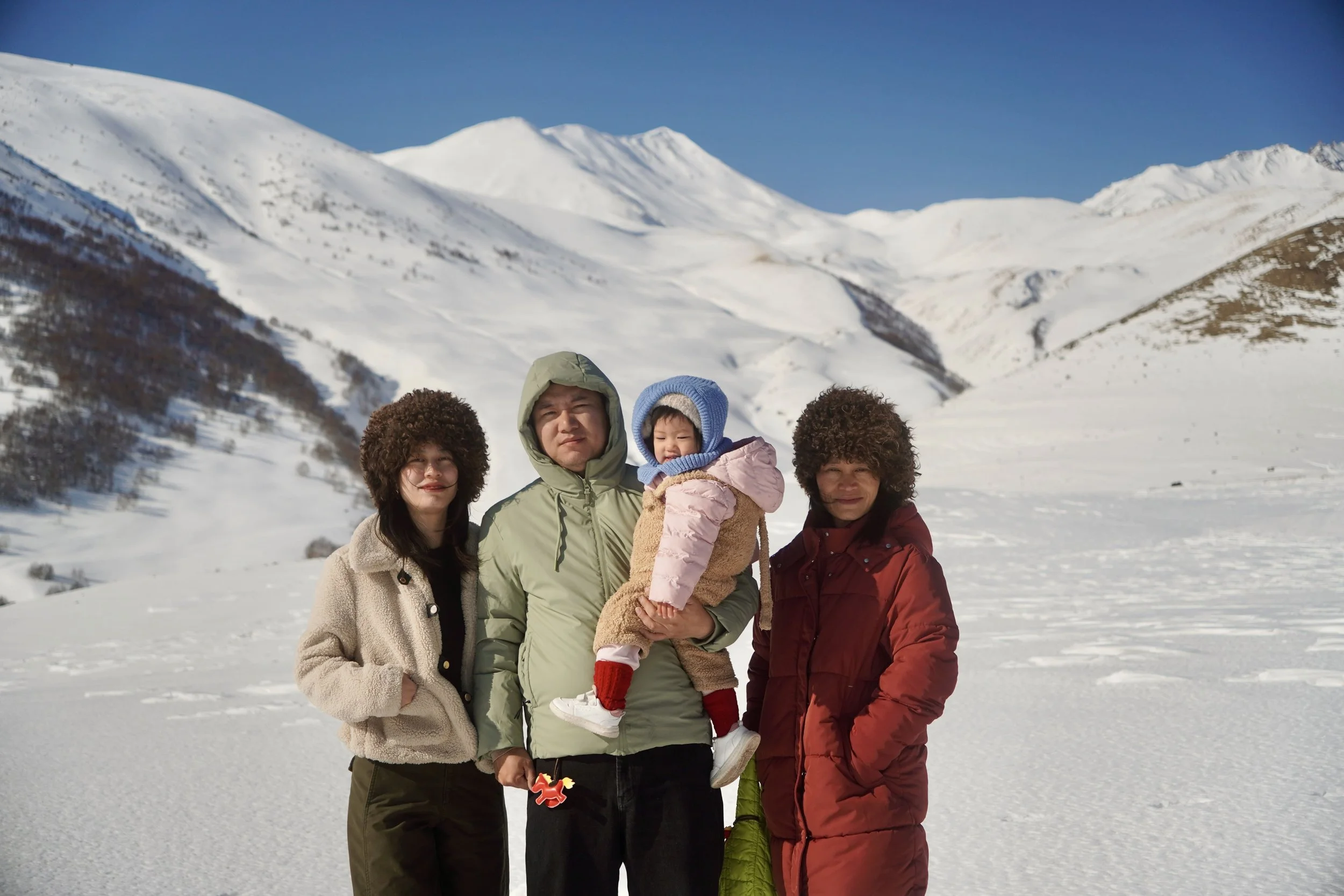 Group of four people, including a child, standing in snowy mountain landscape during daytime, dressed in winter clothing.