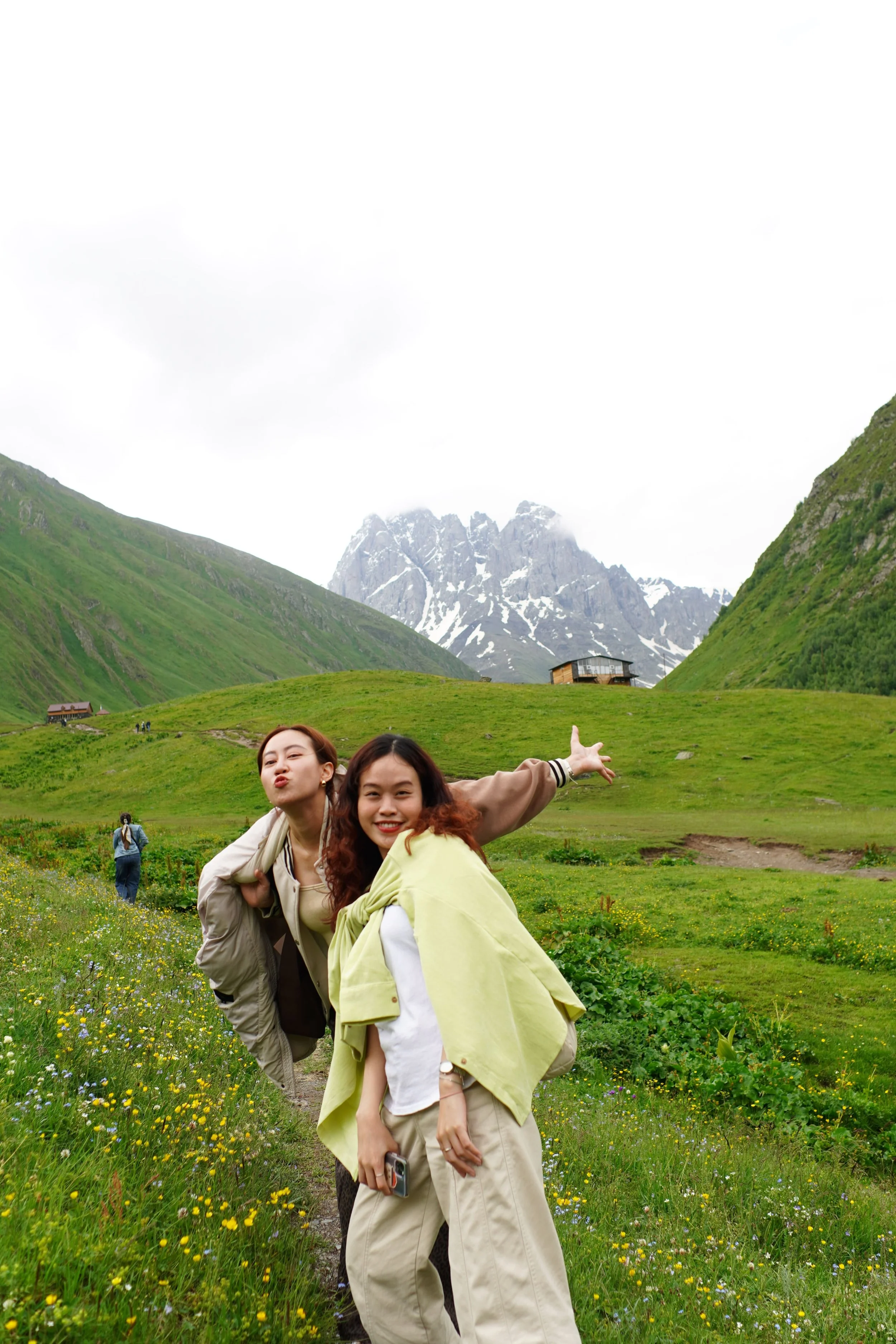 Two women are posing and smiling in a green meadow with mountains and snow in the background. One woman is making a gesture with her arm extended, pointing across the landscape.