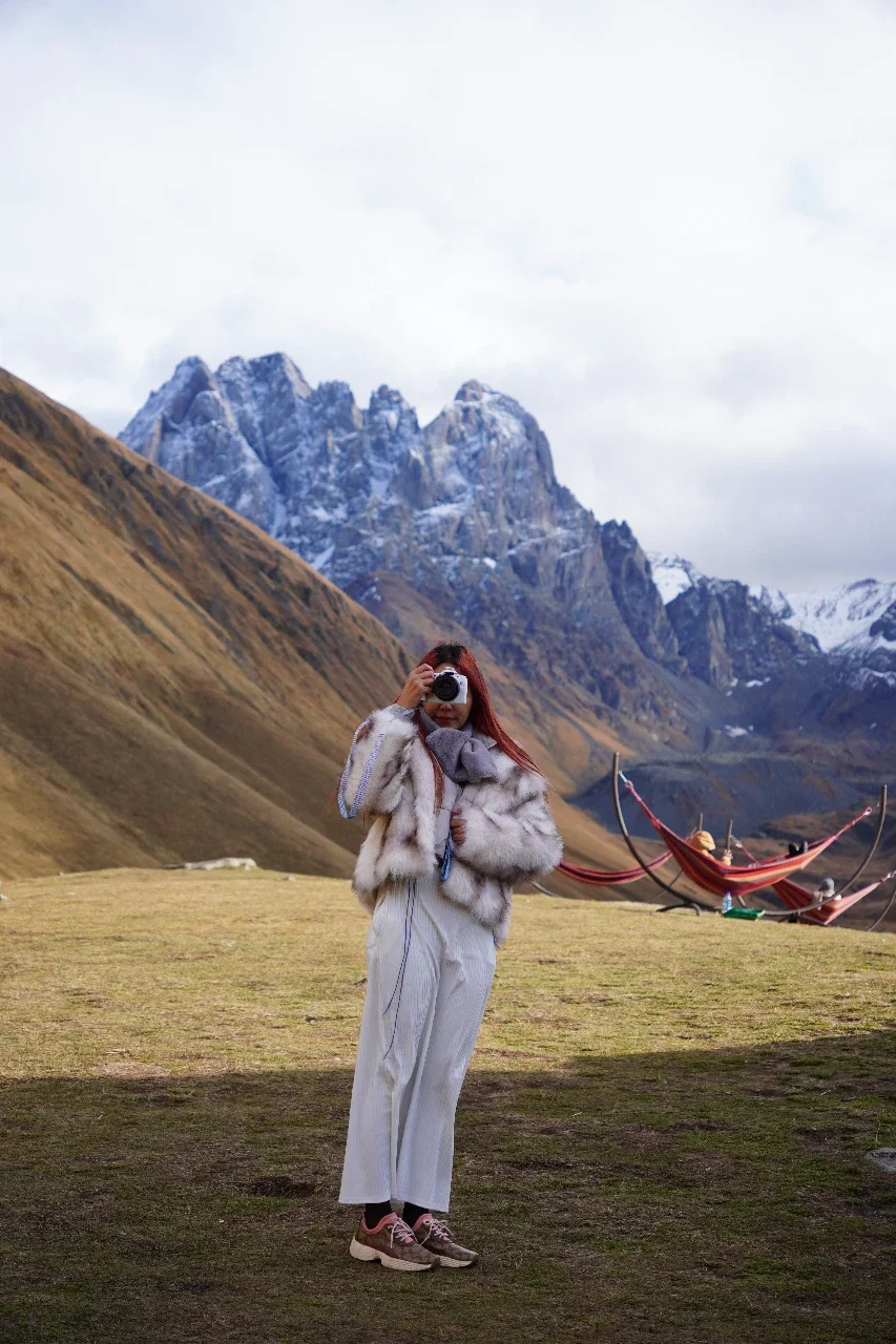 Woman in a fur coat taking a photo with a camera in a mountainous landscape with snow-capped peaks and a hammock in the background.