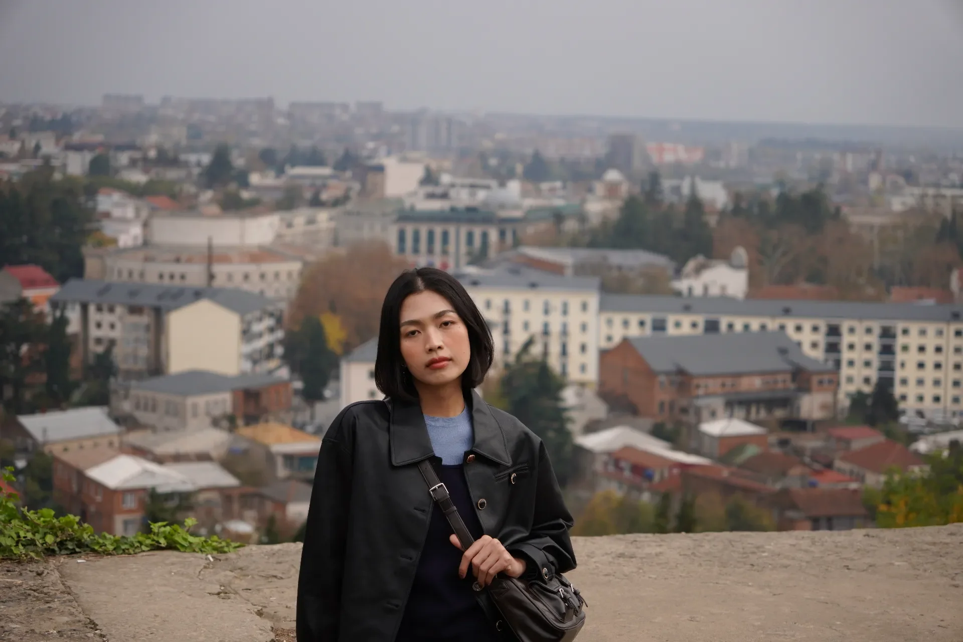 A young woman with shoulder-length dark hair standing outdoors on a concrete surface, with a cityscape of buildings and trees in the background on a cloudy day.