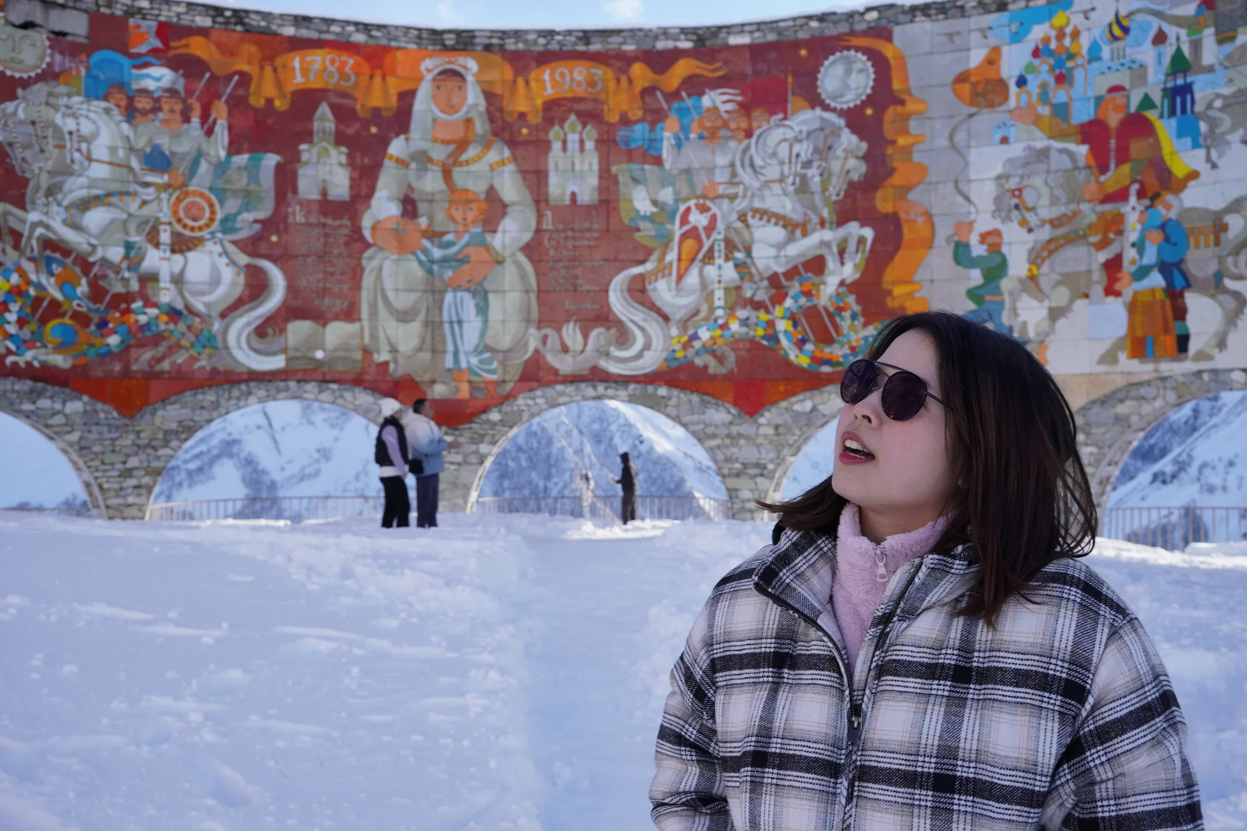 Woman with dark hair, wearing sunglasses, a plaid jacket, and a pink scarf, standing in snowy landscape with a colorful mural depicting historical or cultural scenes in the background.