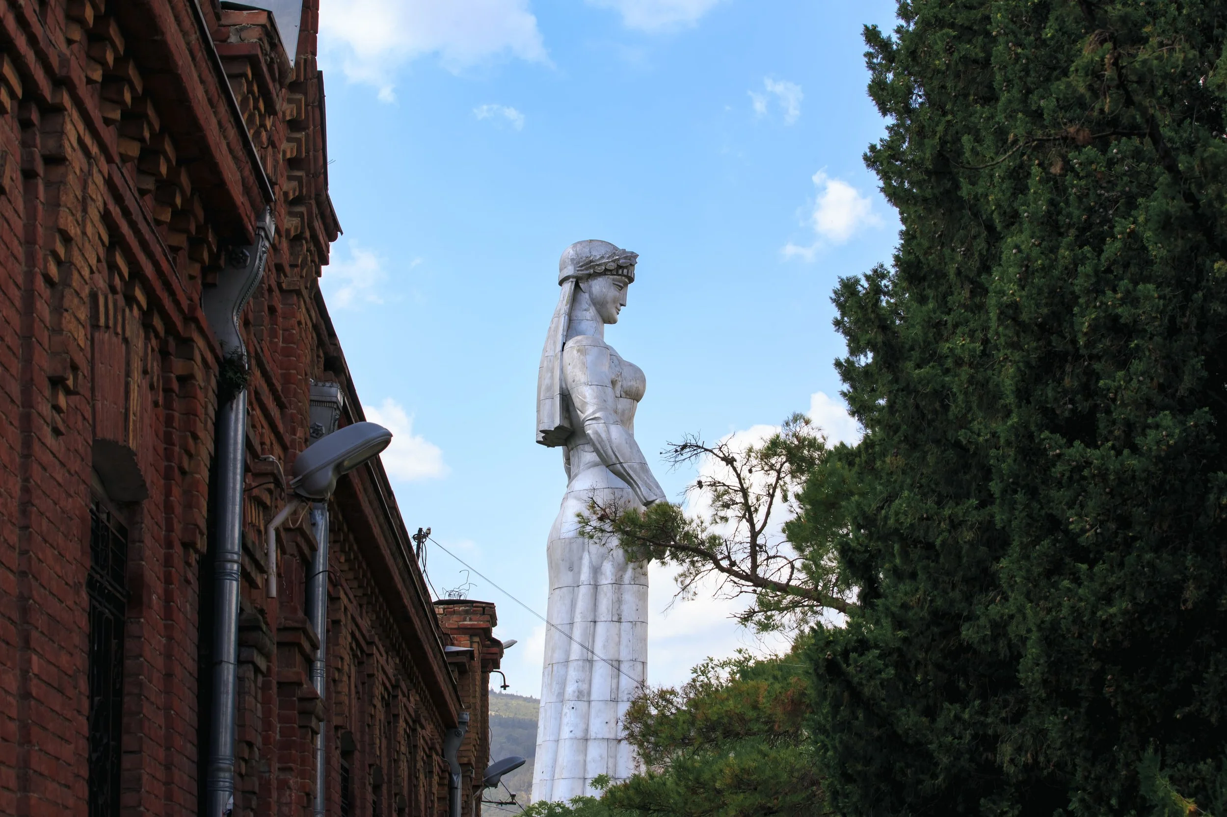 A large white statue of a woman with long hair stands among green trees, with a brick building on the left and a bright blue sky with clouds above.